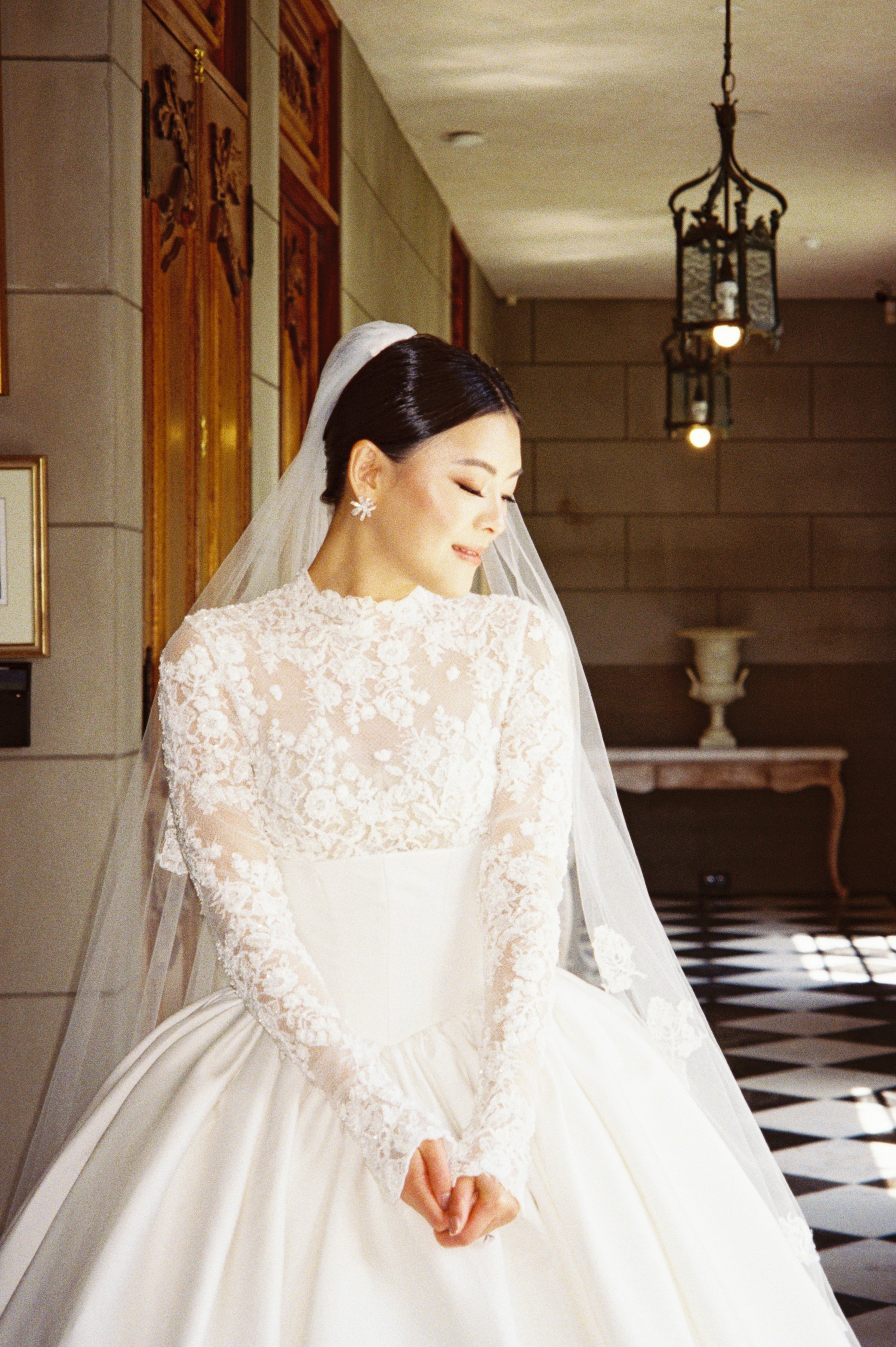 A bride in a white wedding gown with lace sleeves and a veil, standing indoors with ornate wooden wall decorations and hanging lantern lights.