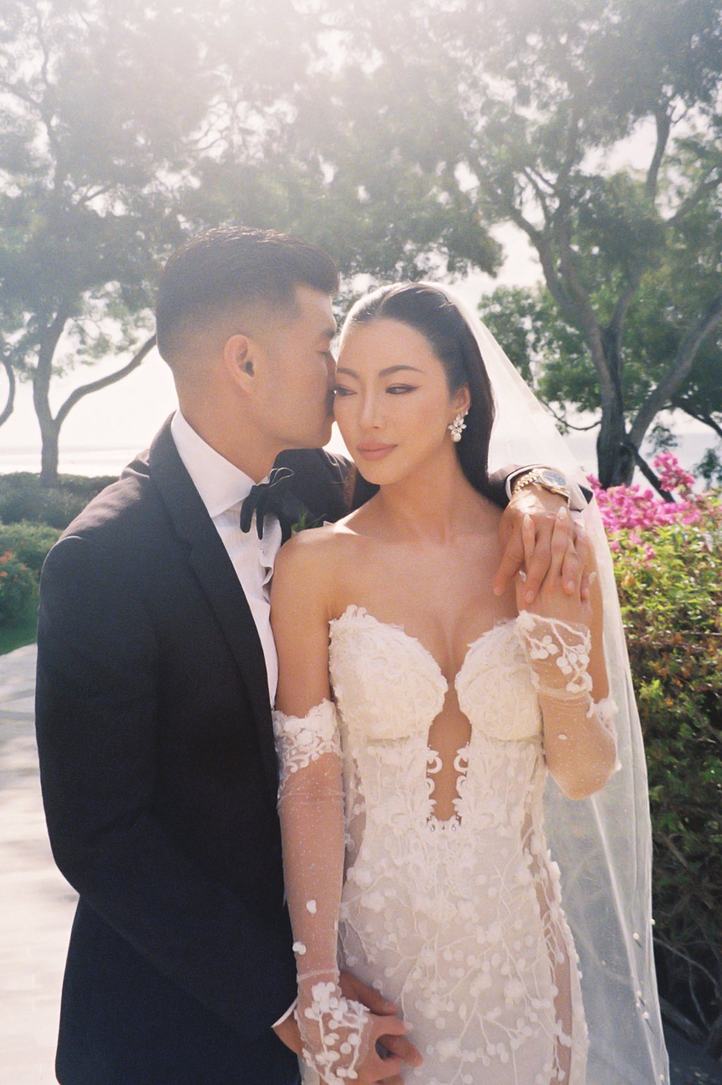 A groom in a black tuxedo with a bow tie kisses a bride in a white lace wedding dress with long lace sleeves outdoors, with trees and pink flowers in the background.