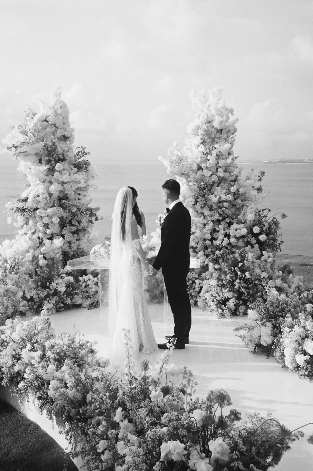 A black and white photo of a wedding ceremony with a bride and groom holding hands, standing in front of floral arrangements near a body of water.
