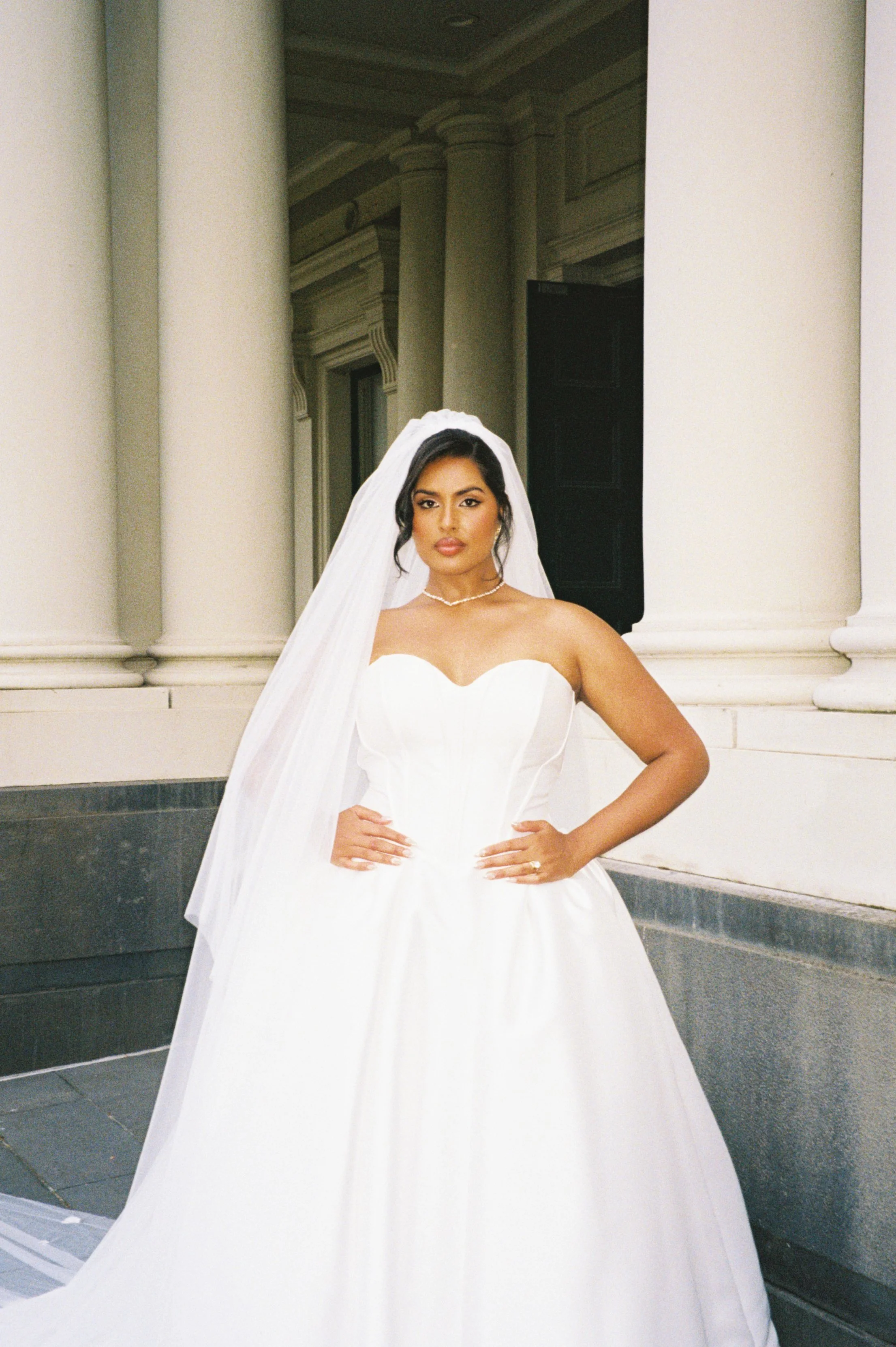 A woman in a white wedding dress standing outdoors in front of large columns.