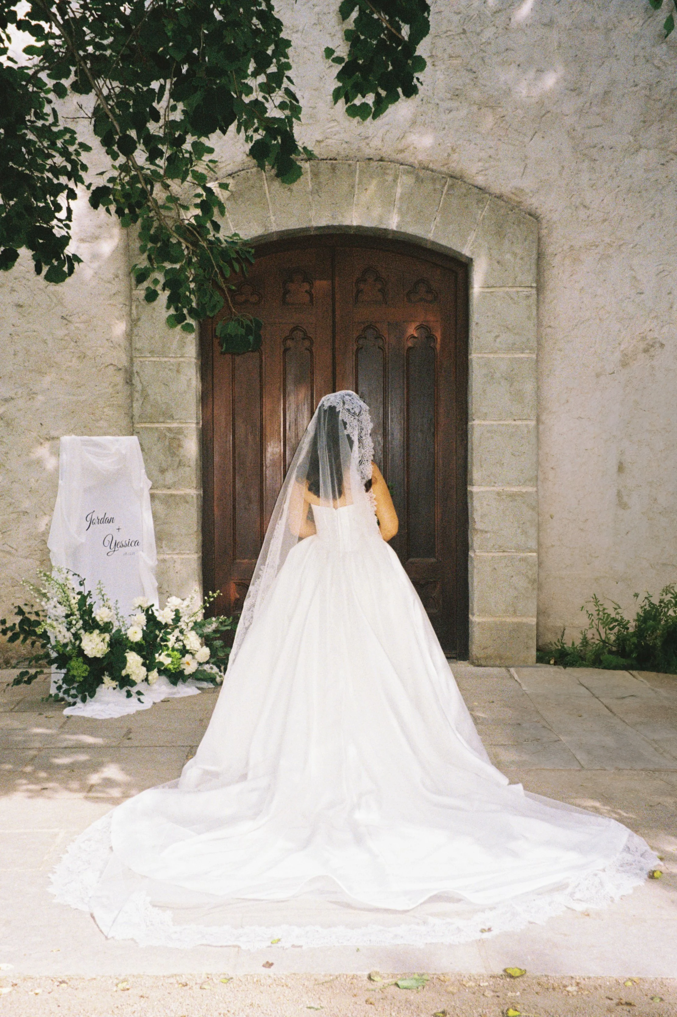 A bride in a white wedding gown and veil standing in front of a wooden door, with floral arrangements and a sign nearby that reads 'Jordan + Jessica.'