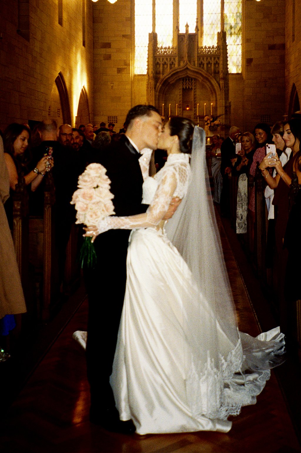 A bride and groom share a kiss in a church during their wedding ceremony, surrounded by guests taking photos.