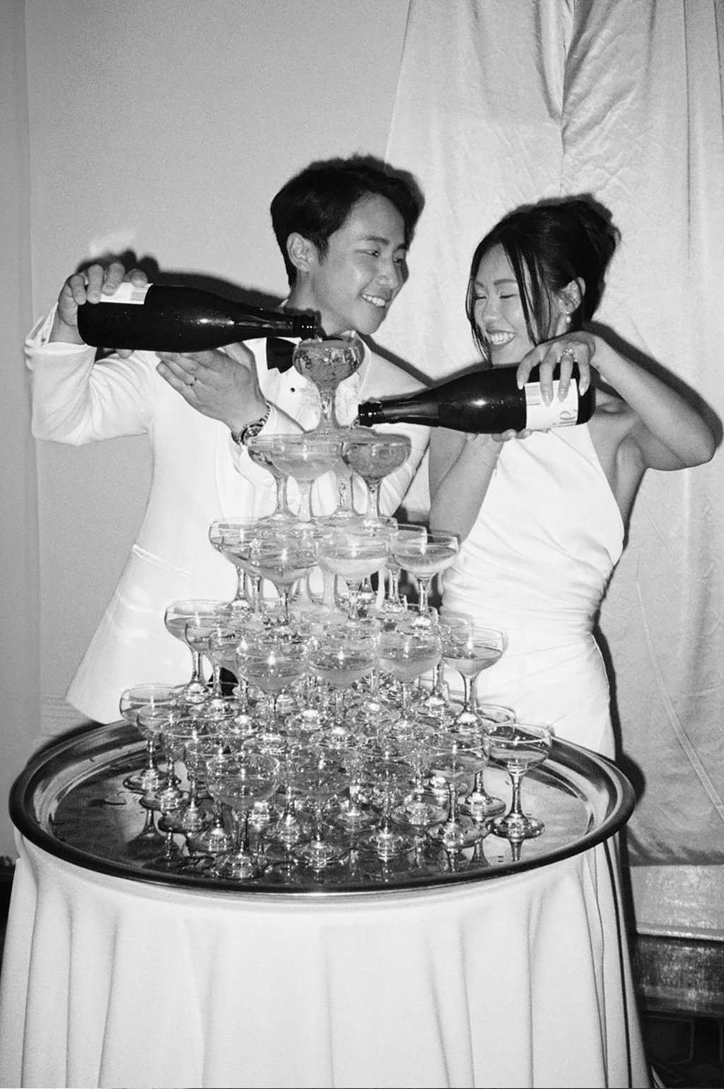 A black-and-white photo of a man and woman pouring champagne into a pyramid of glasses at a celebration or wedding.
