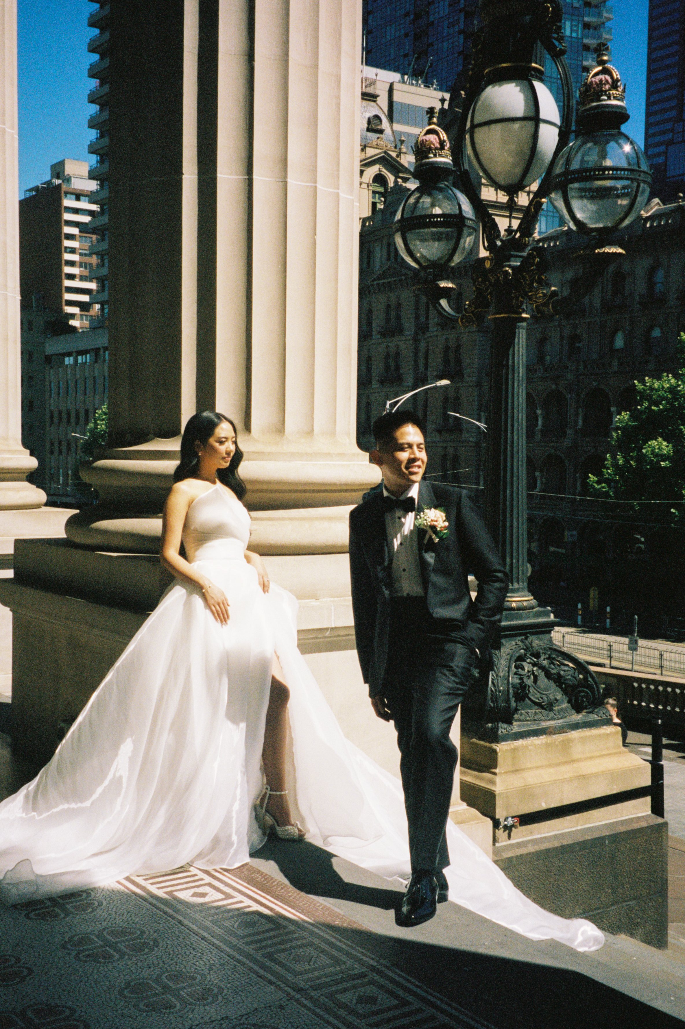 Bride in a white wedding gown and groom in a tuxedo posing outside a historic building with large columns, a streetlight, and a cityscape in the background.