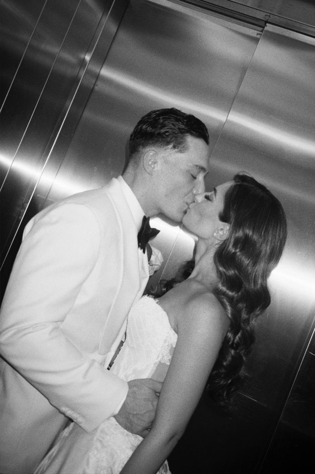 A black-and-white photo of a bride and groom kissing in an elevator, with the groom wearing a tuxedo and the bride in a strapless wedding dress with wavy hair.