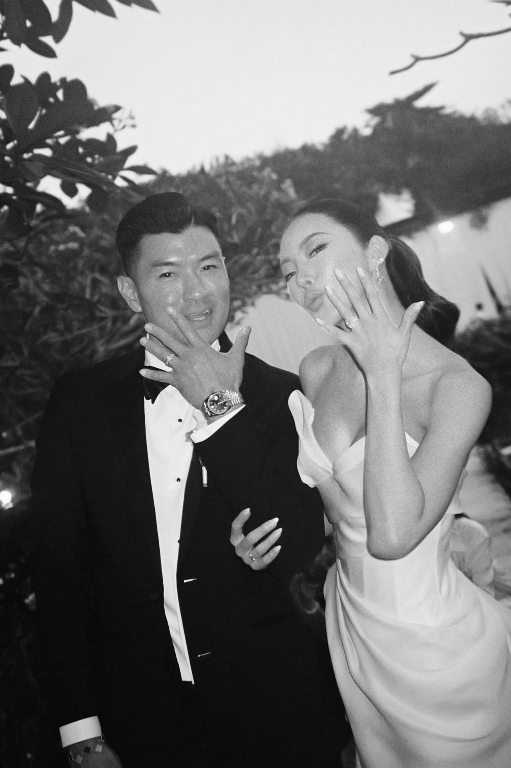 Black and white photo of a man and woman in formal attire, showing off rings on their fingers, smiling, outdoors with trees in the background.