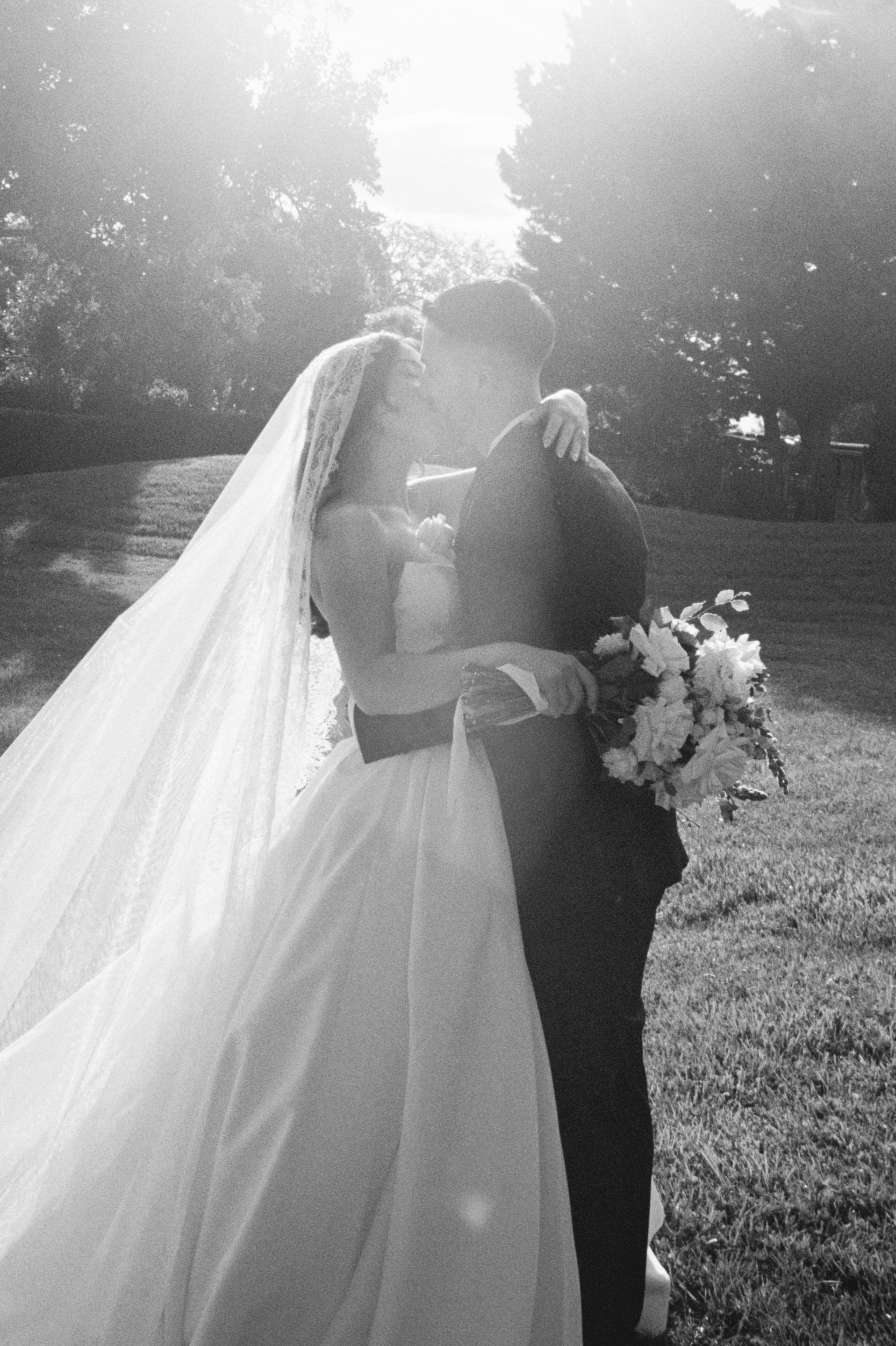 A bride and groom sharing a kiss outdoors, with the bride holding a bouquet of flowers, in a black and white photo.