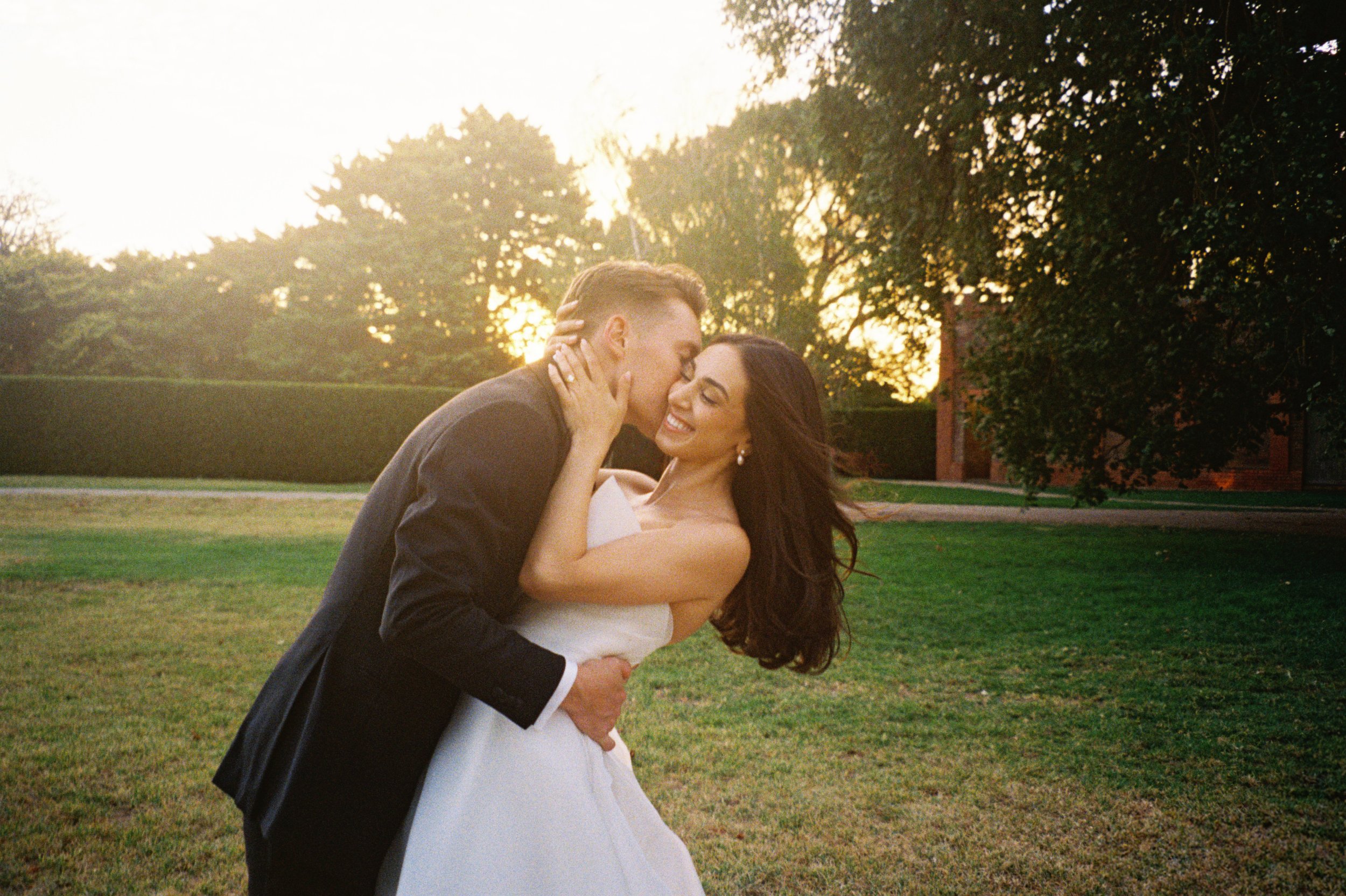 A newlywed couple sharing a kiss outdoors on a grassy field at sunset, surrounded by trees.