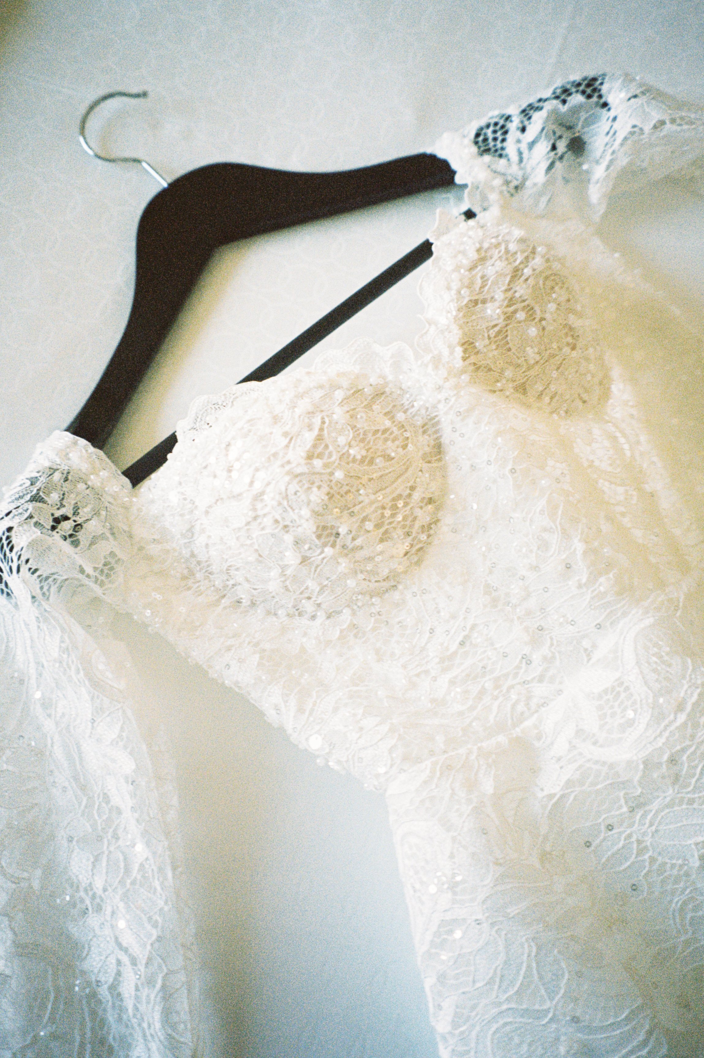 A white lace wedding dress hanging on a dark wooden hanger against a light-colored wall.