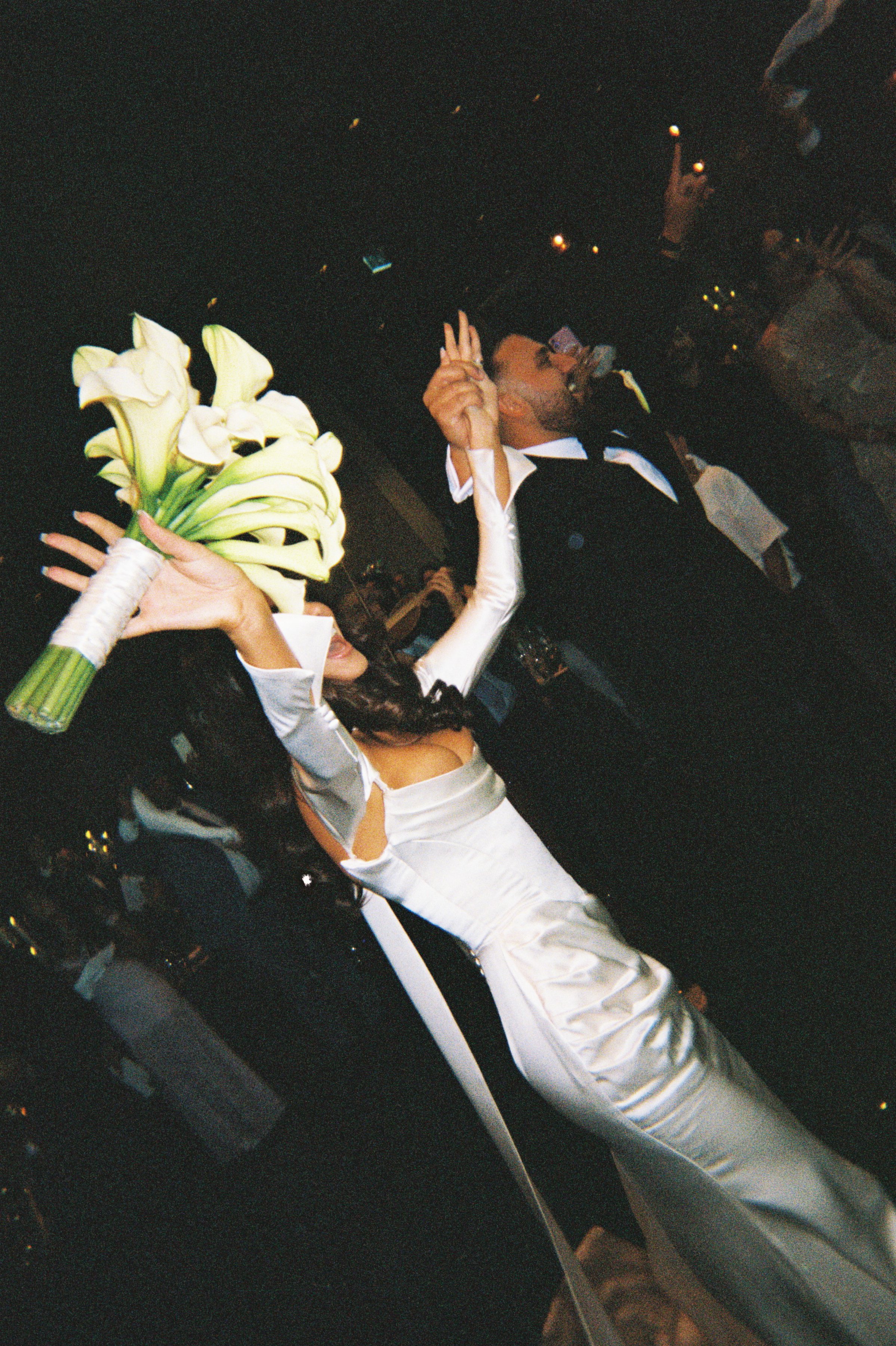 Bride and groom dancing at their wedding reception, with the bride holding a bouquet of lilies and making a peace sign.