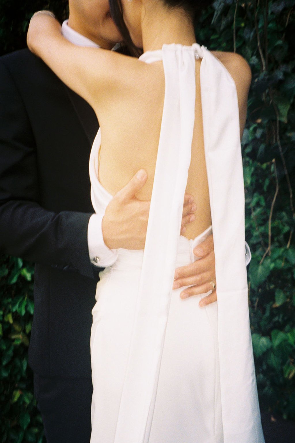 Close-up of a bride in a white wedding dress embracing a groom in a black suit, with a background of green leafy plants.