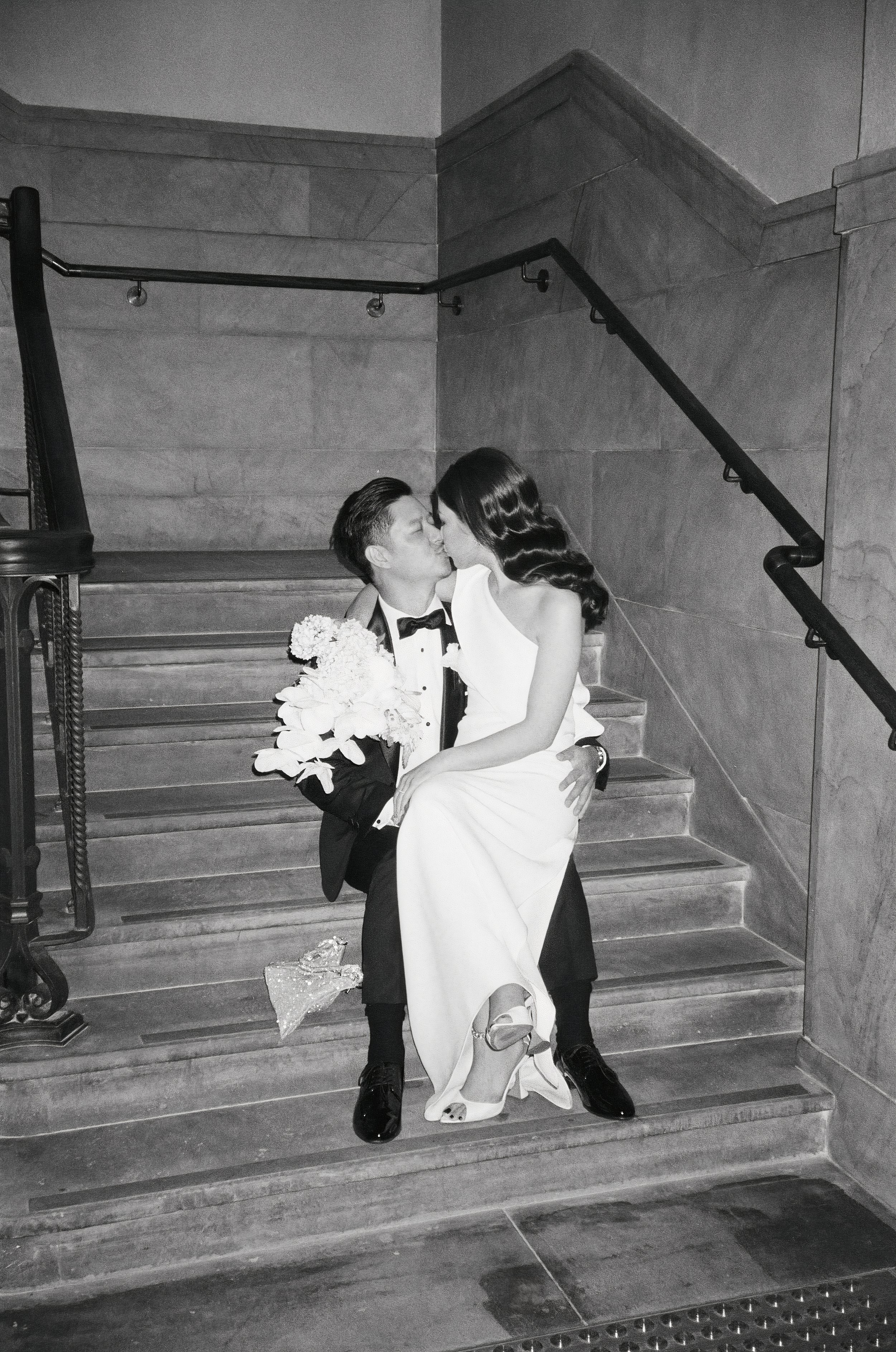 A black-and-white photo of a bride and groom sharing a kiss on a staircase, with the bride sitting on the groom's lap, holding a bouquet of flowers, and wearing a white dress, while the groom is in a tuxedo.