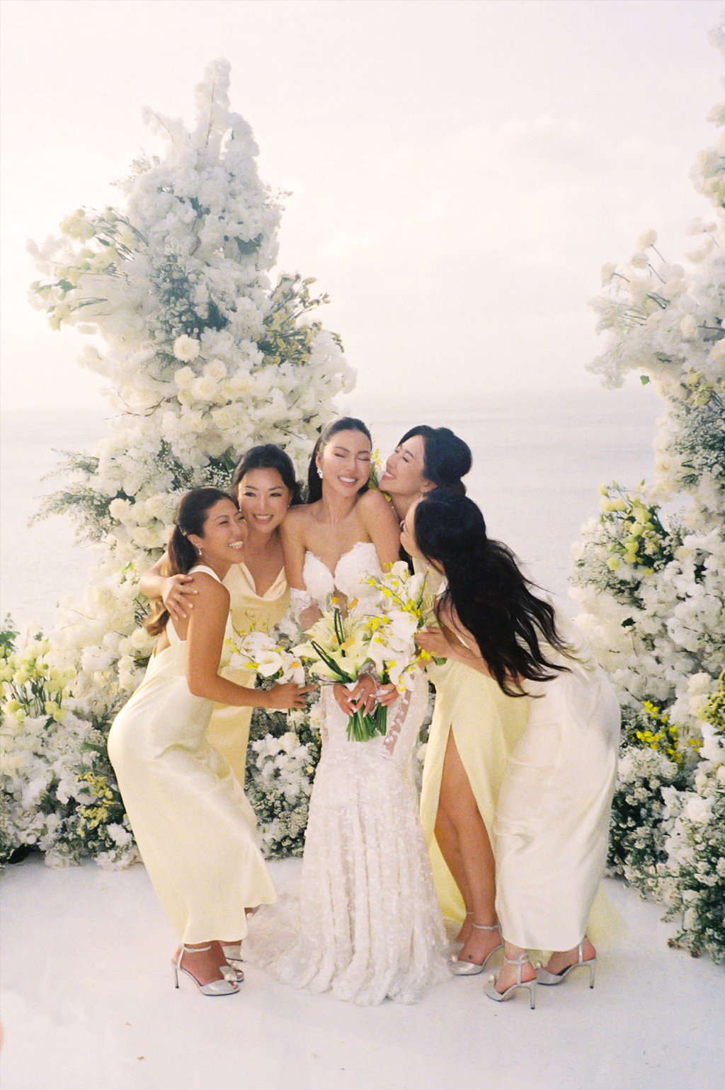 A bride in a white wedding dress holding a bouquet of white flowers, surrounded by four women in yellow dresses, on a floral decorated stage with white flowers.