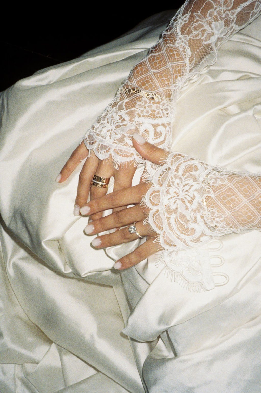 Close-up of hands adorned with rings and jewelry, resting on a white satin fabric, with lace wedding dress sleeves in the background.