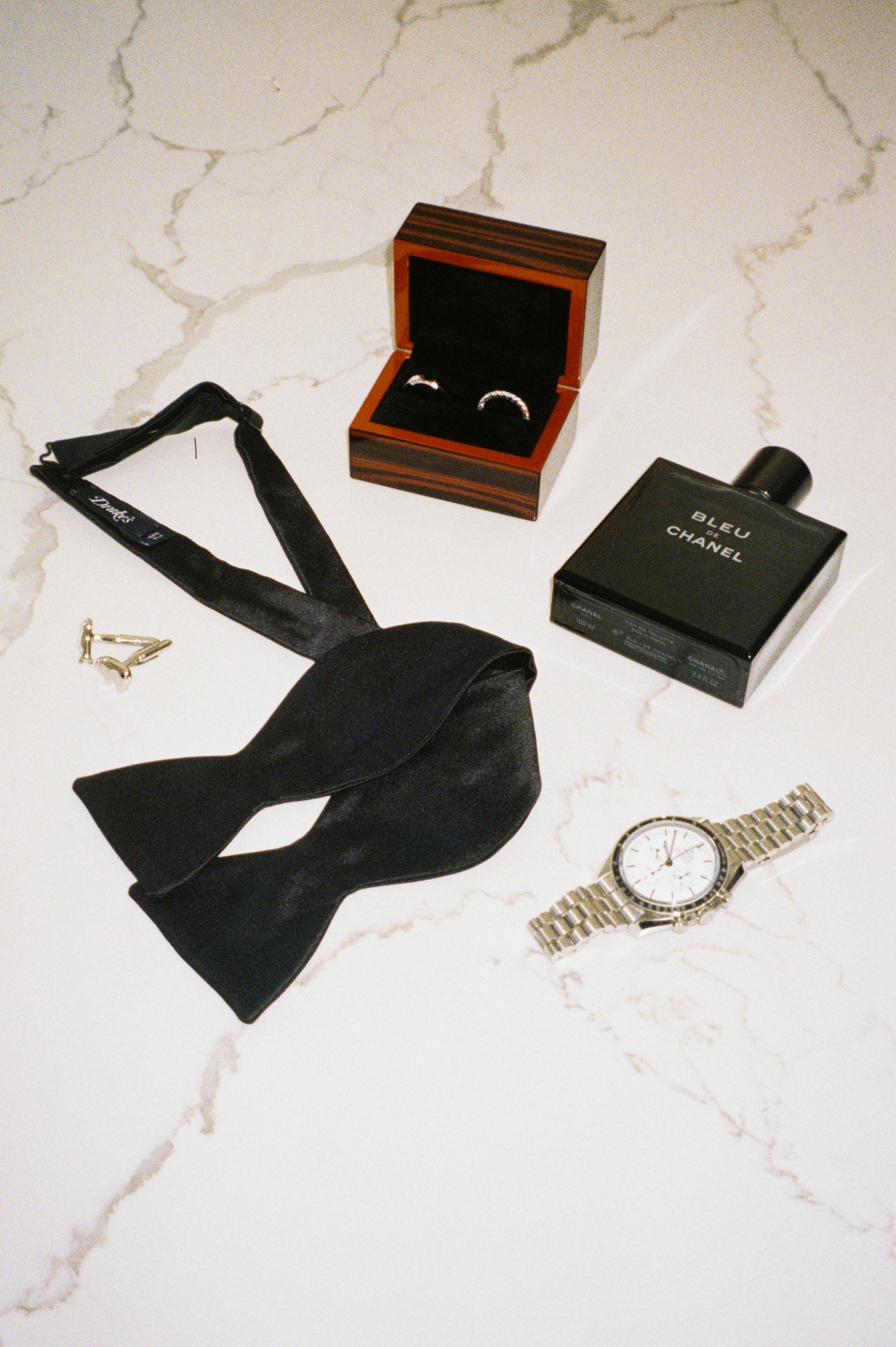 A collection of men's accessories including a silver wristwatch, gold cufflinks, a black leather bow tie, a black tuxedo belt, a ring in a wooden box, a box of Chanel Bleu de Chanel cologne, and a pair of earrings with a black box.