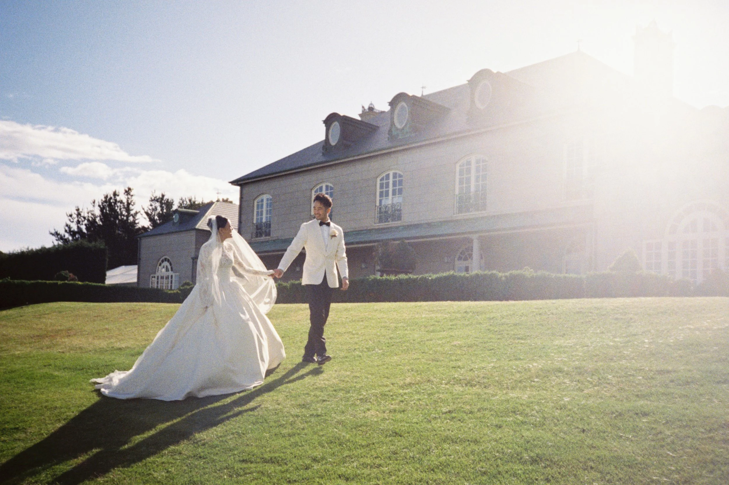 A bride and groom holding hands and walking on a lawn in front of a large house, with sunlight shining brightly from the right side of the image.