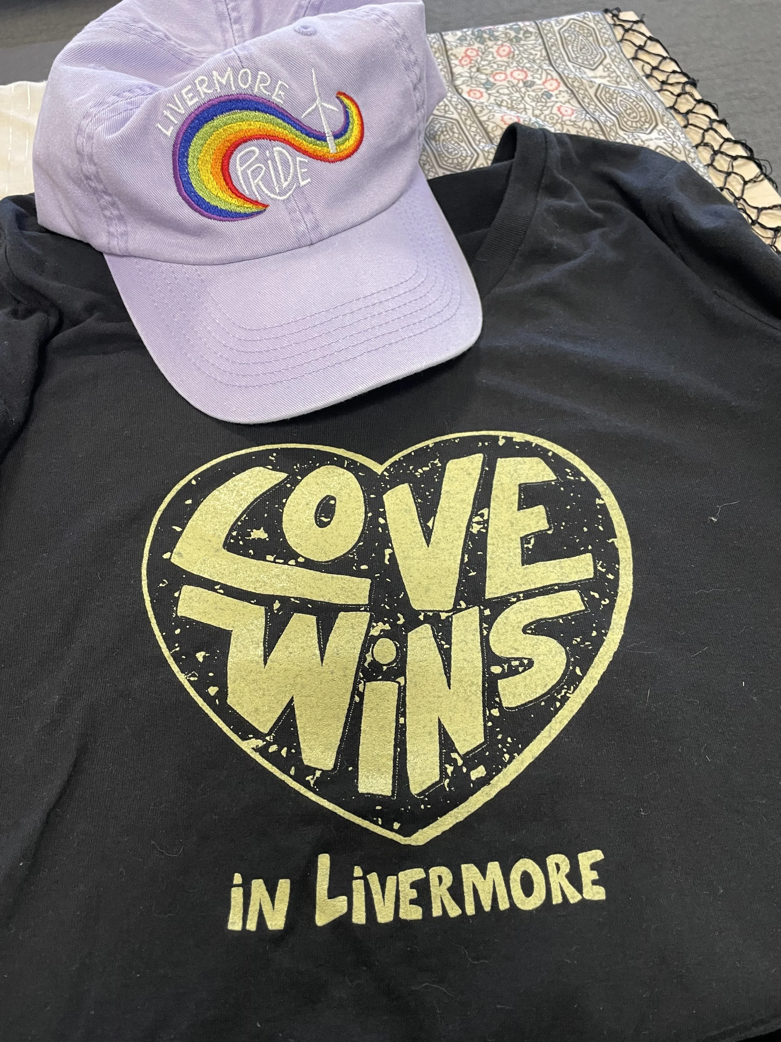 Black t-shirt with lettering "love wins in livermore" and a lavendar baseball hat with Livemore Pride logo that was hand lettered and drawn, embroidered on the front.