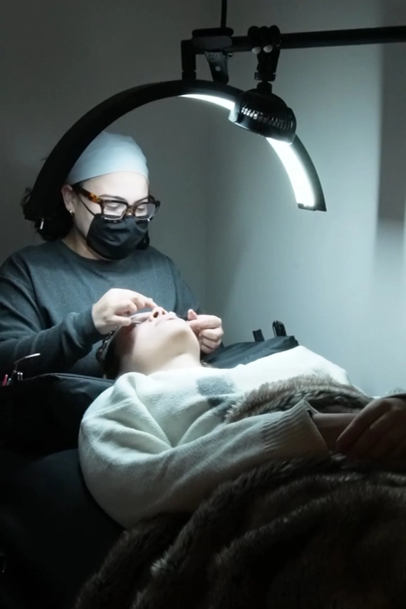 A woman receiving an eyelash extension treatment by a technician under a bright lamp in a salon.