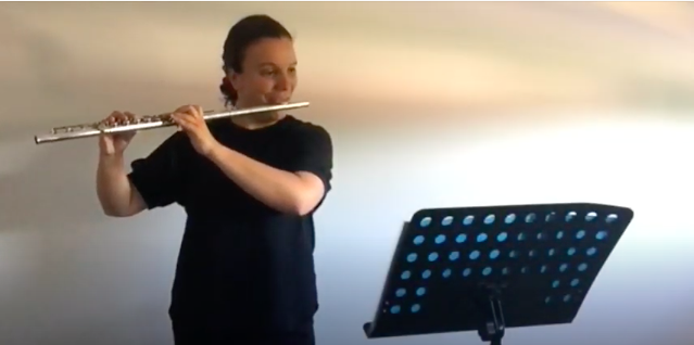 Kelsy plays the flute, reading from a music stand against a white wall.