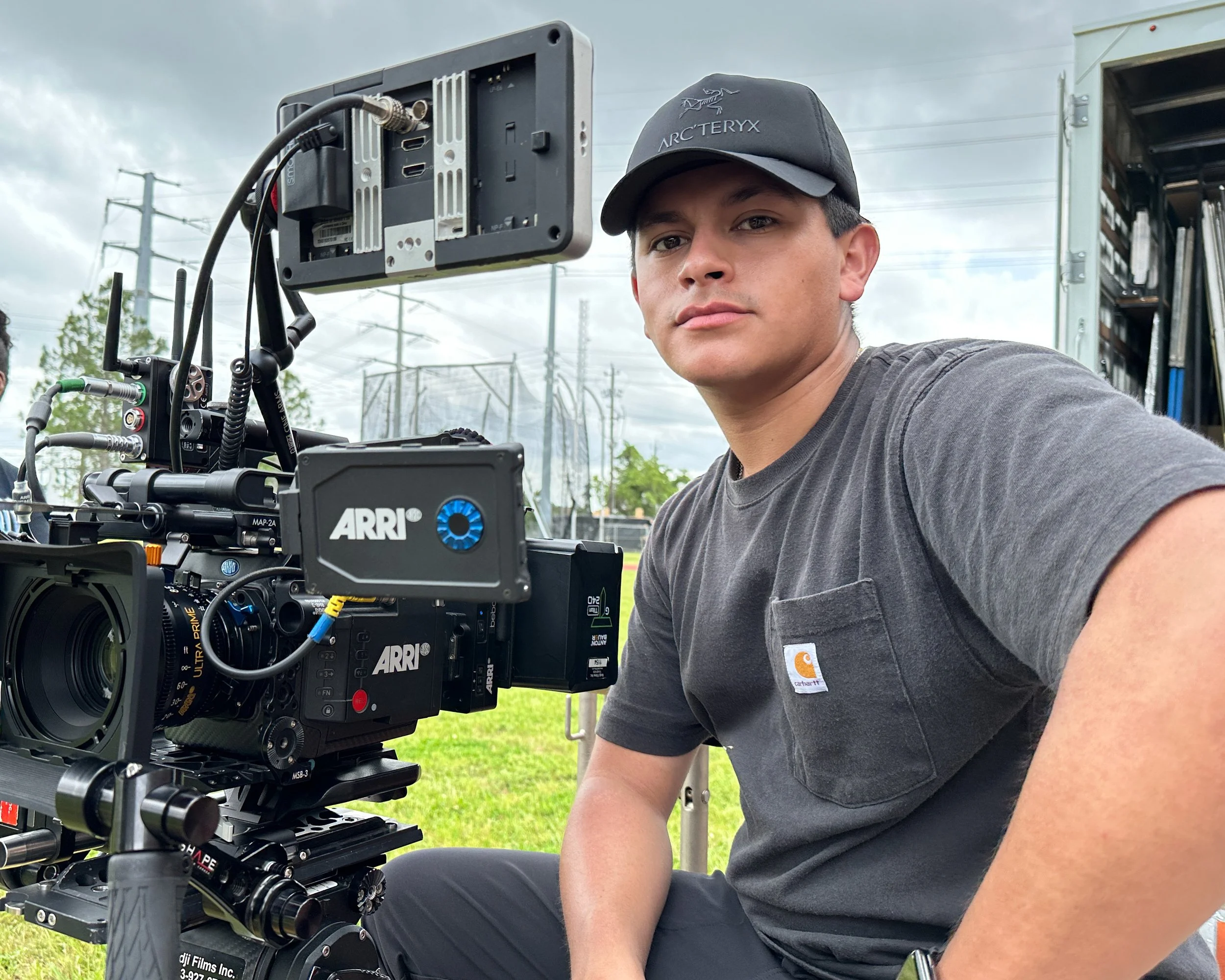 Young man operating professional film camera outdoors on grassy field, cloudy sky in background.