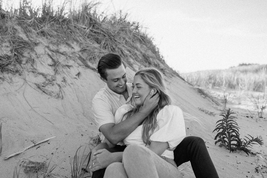 Engaged couple smiling and laughing while boyfriend holds girlfriends face sitting on the beach