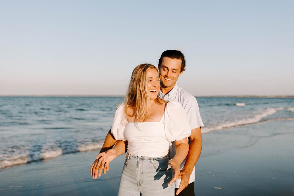 Boyfriend grabbing girlfriend from behind while they laugh and smile on Plum island beach for their beach engagement photoshoot