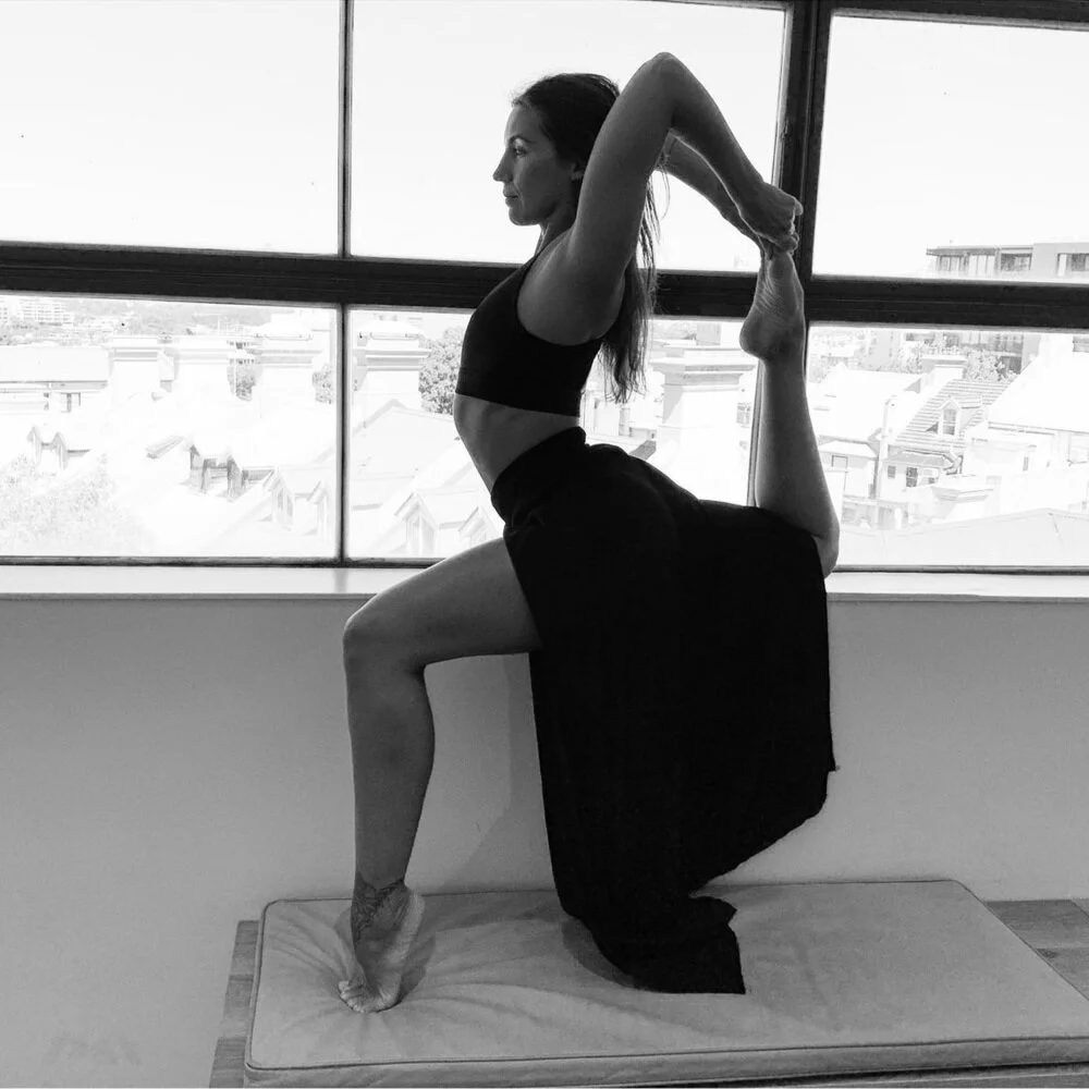 A woman in a yoga pose, balancing on her left foot with her right foot raised behind her, held by both hands, near a window showing a cityscape.
