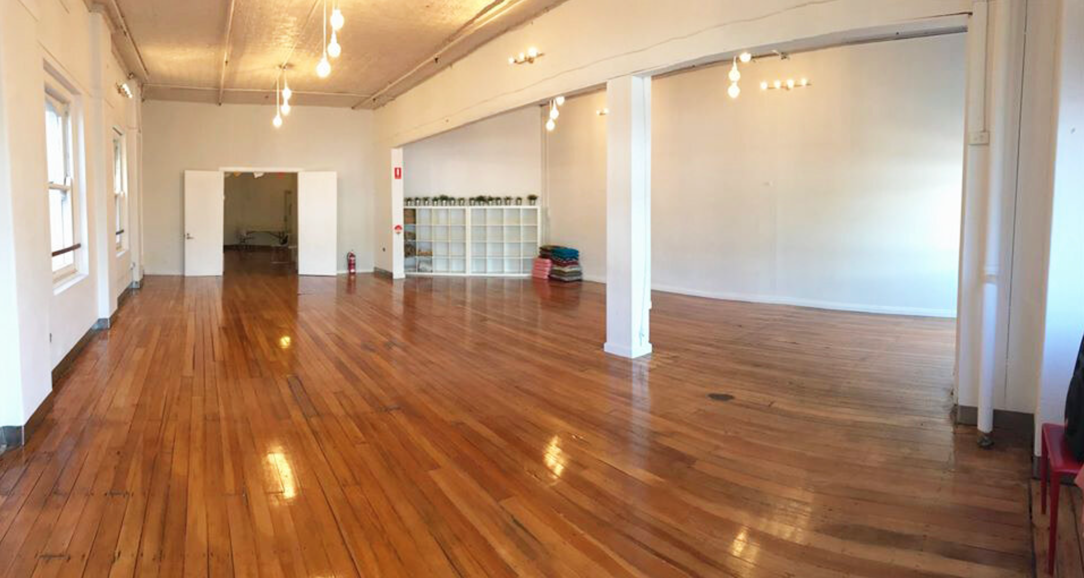 Empty spacious room with polished hardwood floors, white walls, multiple windows, overhead lights, and a white door leading to another area.