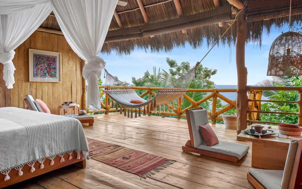 A rustic bedroom with a bed covered in white fabric and pink pillows, a wooden deck with a hammock facing lush greenery and the ocean, and cozy chairs with cushions, all within a thatched roof structure.