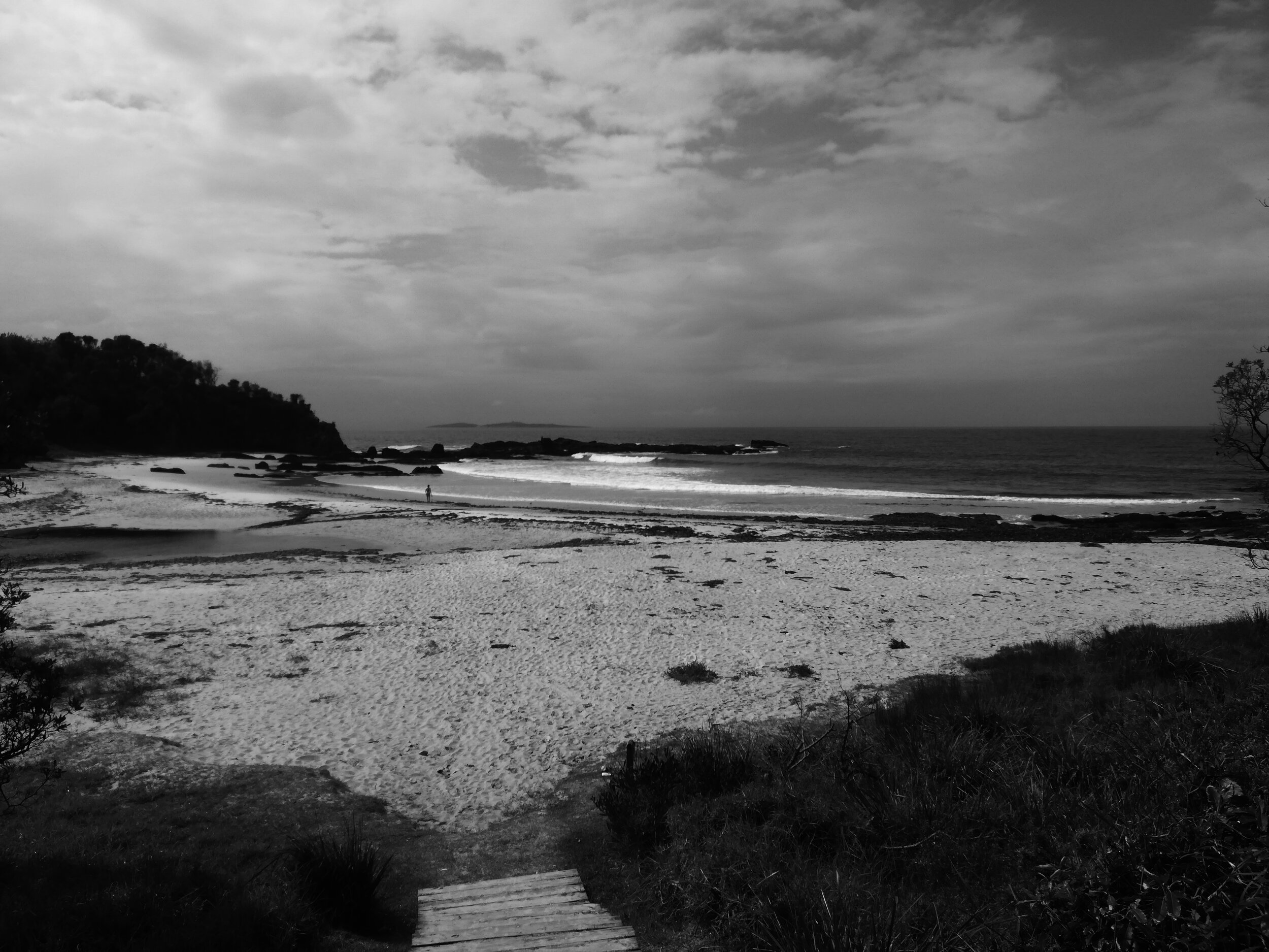 Black and white photo looking down from a few steps at the a cove shaped beach and out across a few small waves to an expansive ocean.