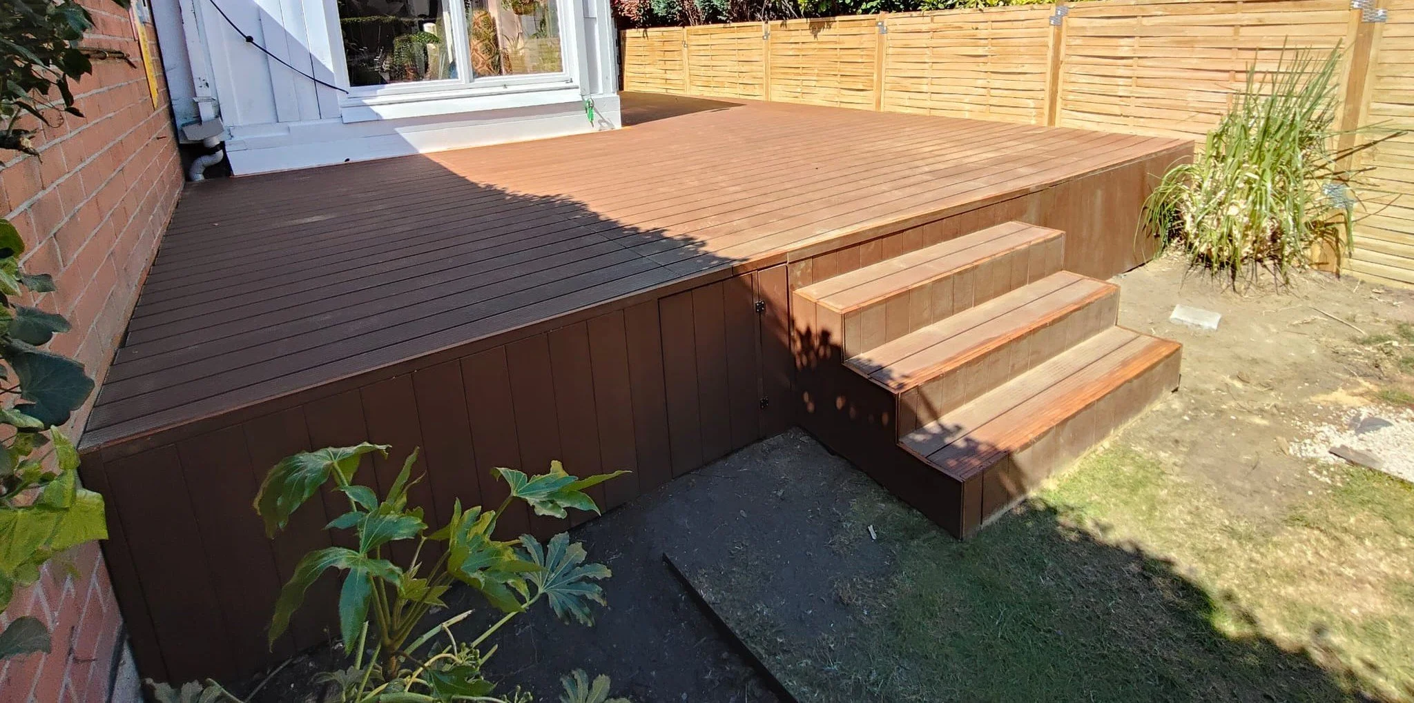 A newly built wooden deck with stairs extends from a house with a white door and window, surrounded by a brick wall on one side and a wooden fence in the background, with some plants and grass nearby.