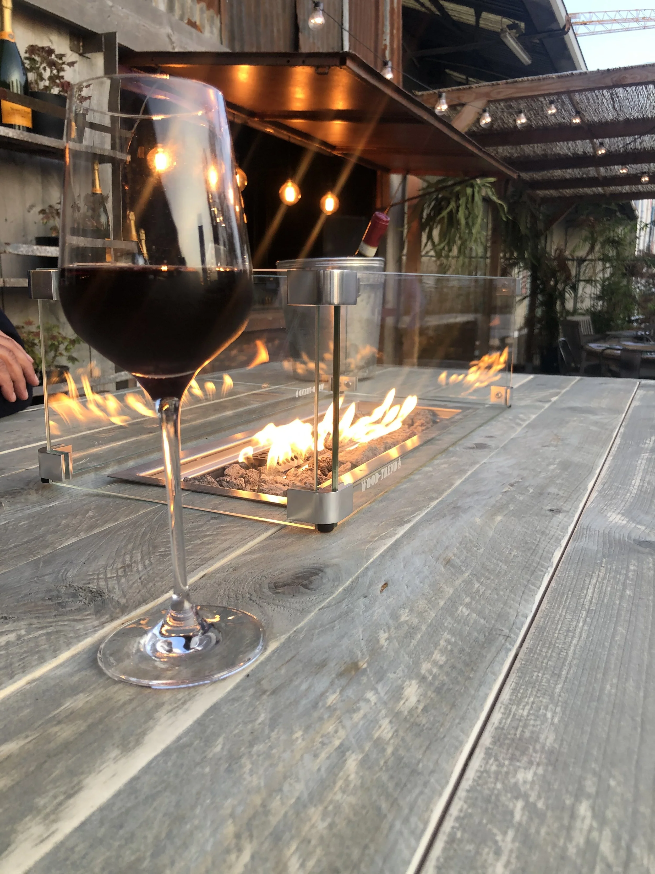 Glass of red wine on a wooden table in an outdoor setting with a tabletop fire pit and string lights.