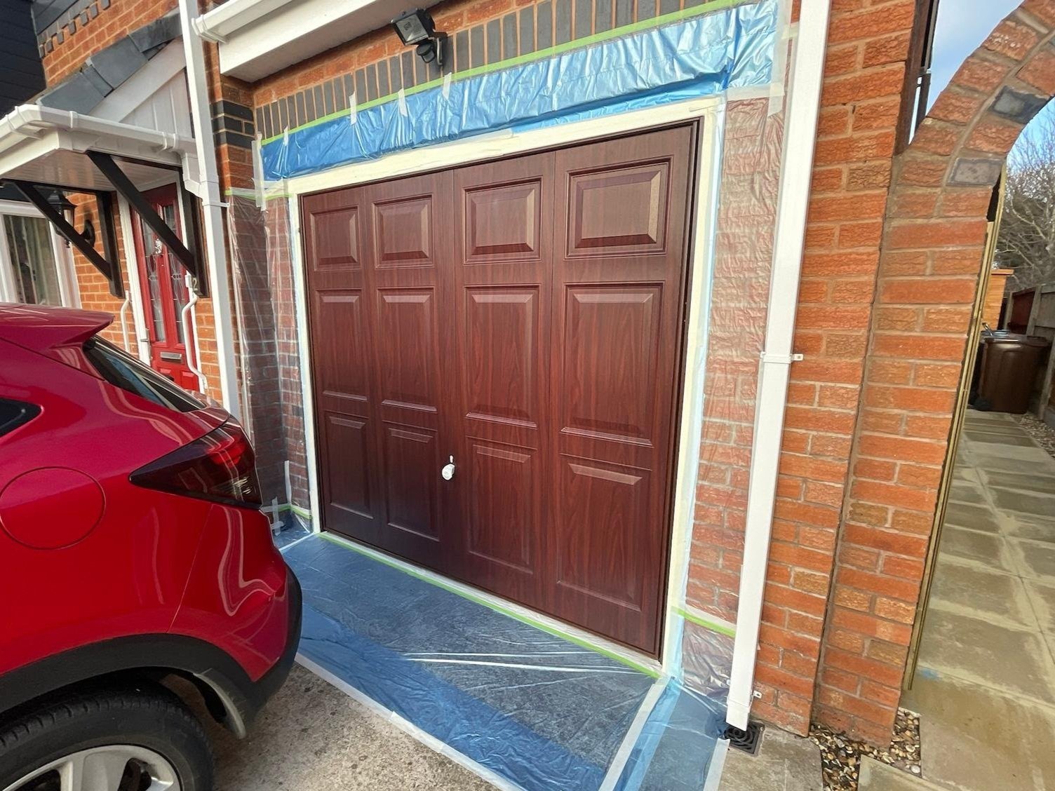 Red car parked in front of a new garage door with masking tape and plastic sheeting around it, indicating it is under installation or painting.