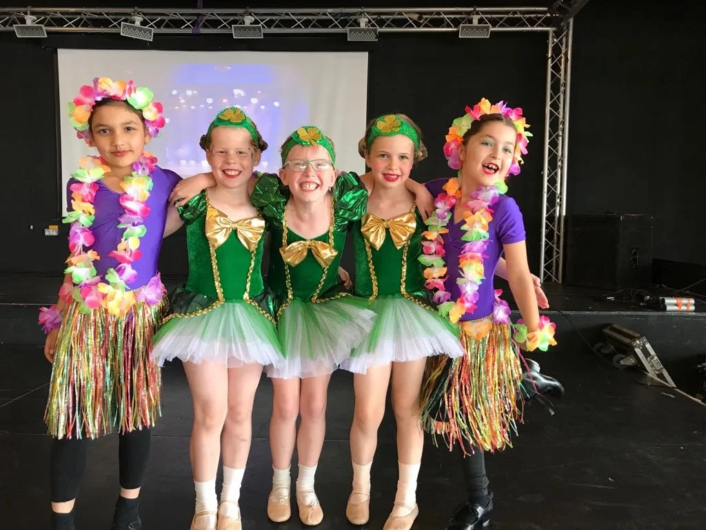 Five young dancers at Just Dance, dressed in vibrant green and gold costumes with floral leis and grass skirts, smiling brightly on stage after a lively and colourful performance.