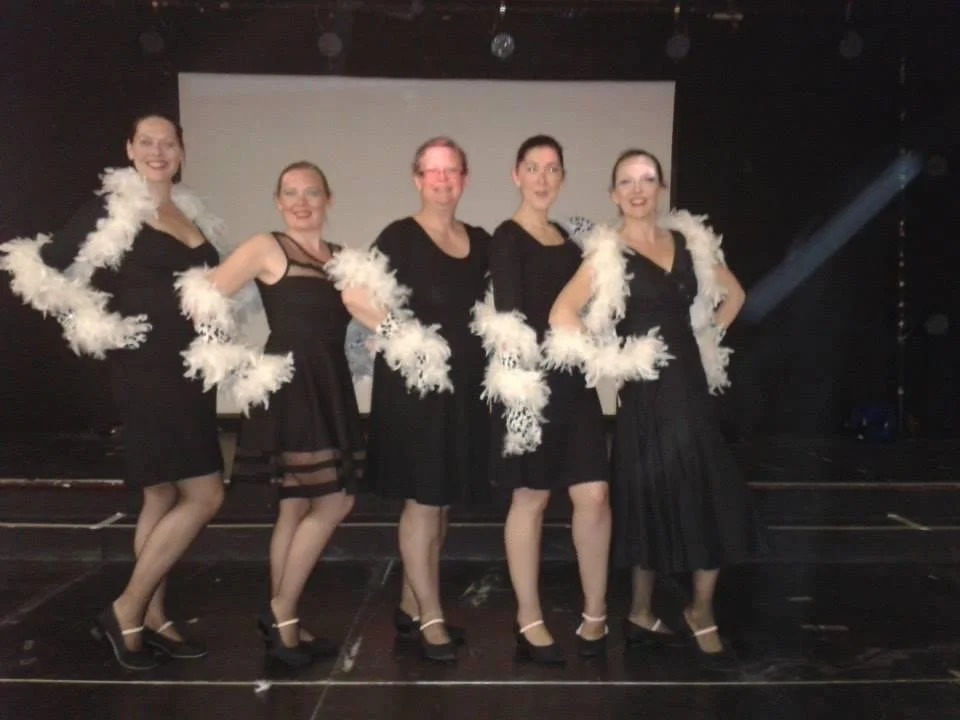 Adult dancers at Just Dance posing in elegant black dresses with feather boas, ready to perform a stylish routine on stage.