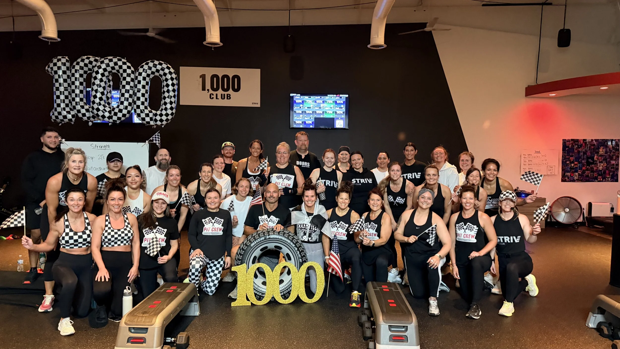 Group of women and men celebrating a 1000-pound club achievement at a fitness gym, with some holding checkered flags and a large "1000" sign, in front of a dark wall with multiple screens.