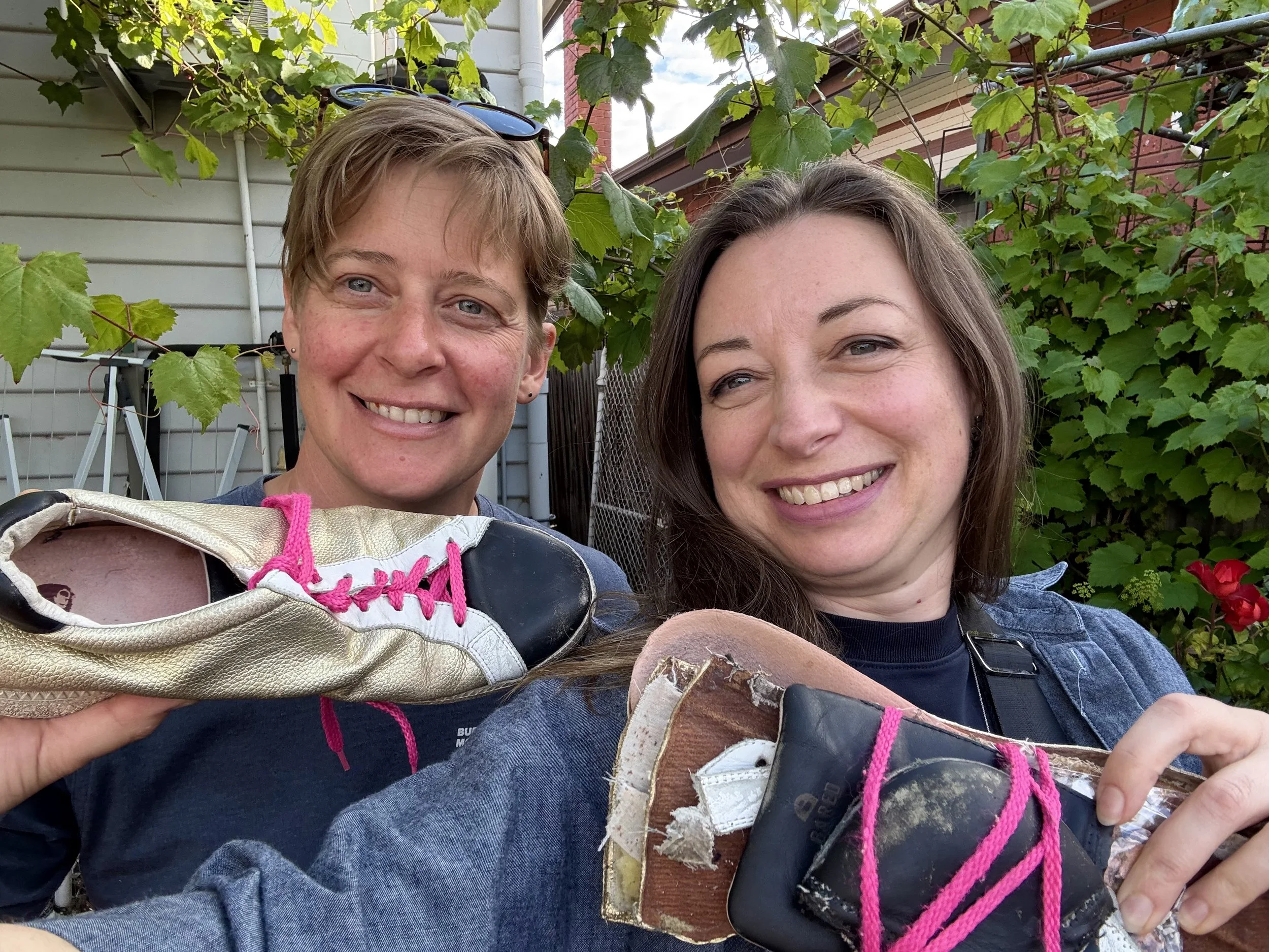 Two smiling women standing in a garden, one holding a gold and black shoe with pink laces, the other holding a bundle of deconstructed shoe materials.