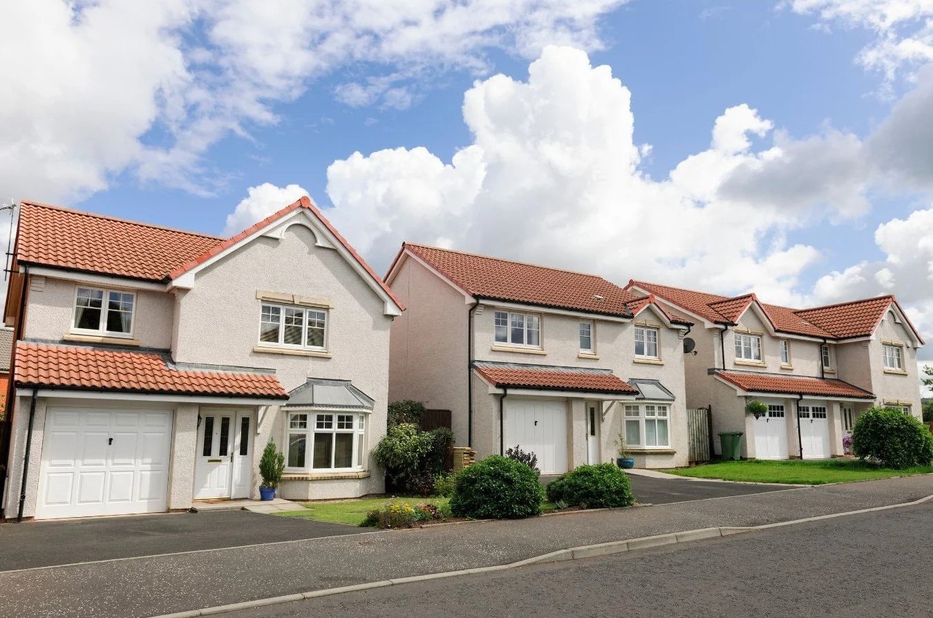 A row of modern suburban houses with white walls and red tiled roofs, neighboring a paved street under a partly cloudy sky.