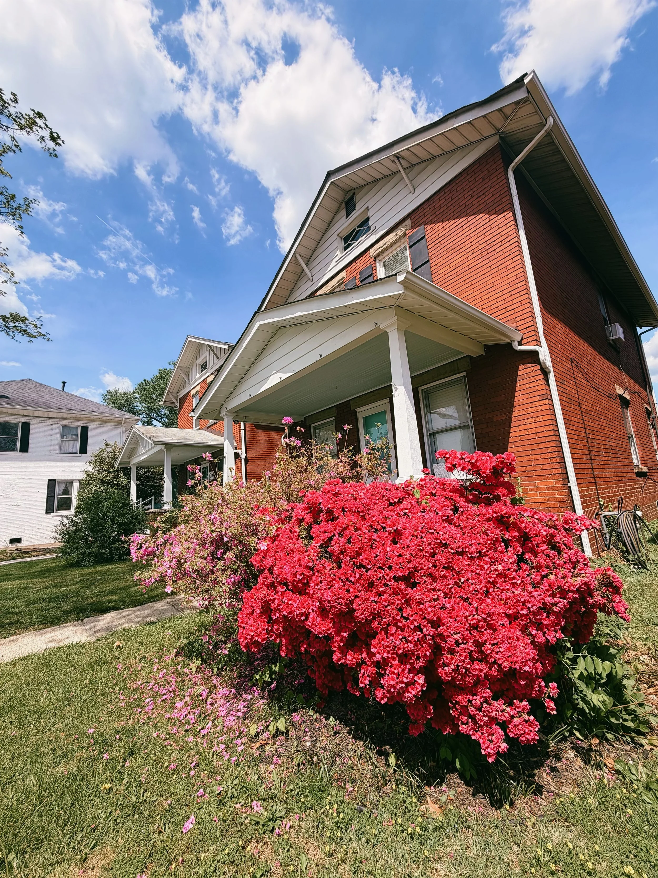 A red brick house with white trim and a porch, surrounded by pink and purple flowers, under a blue sky with white clouds.