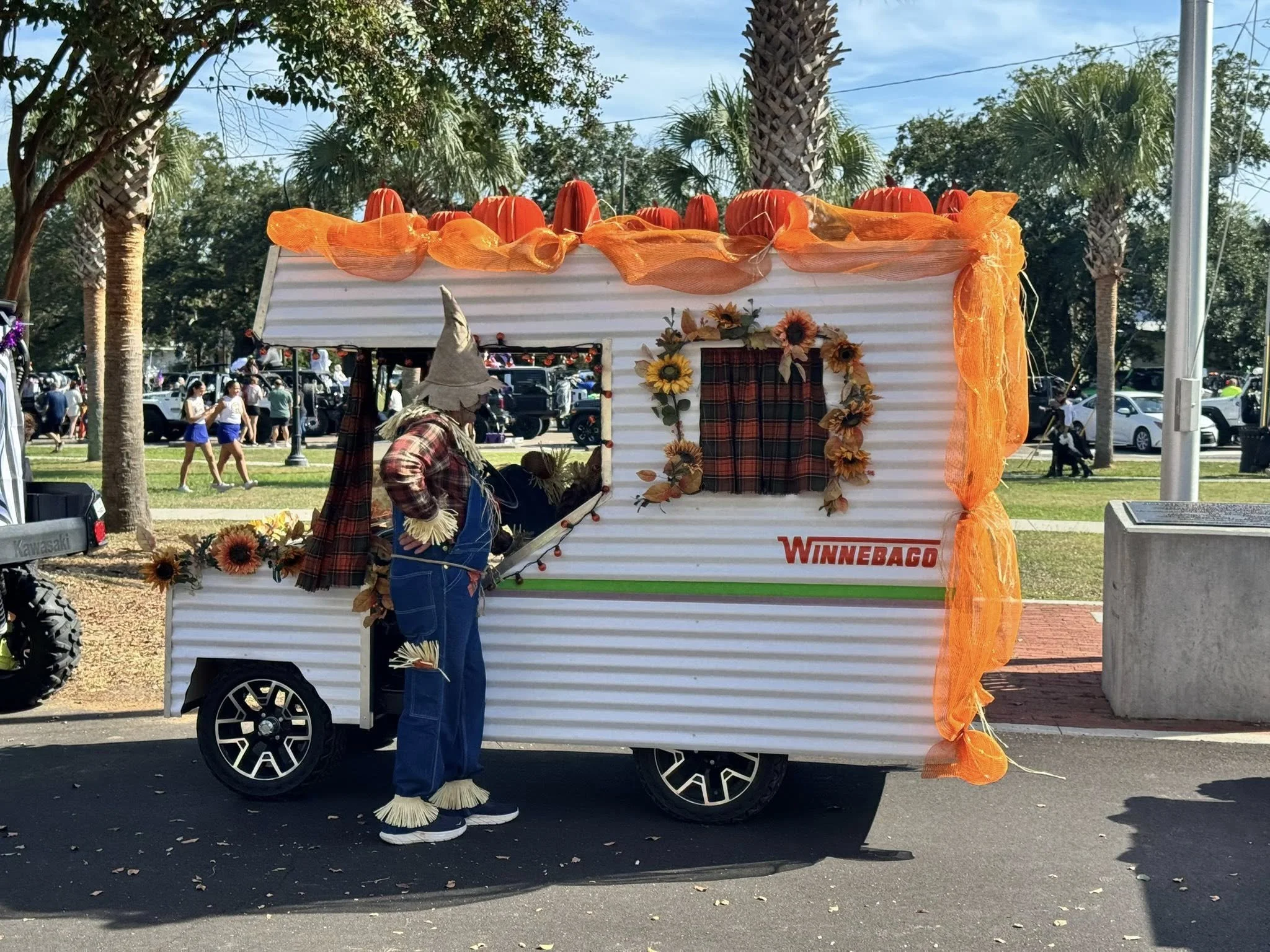 A food trailer decorated with orange fabric, sunflowers, and fall leaves, with a person wearing a scarecrow-themed costume in front of it at an outdoor event, surrounded by palm trees and parked cars.
