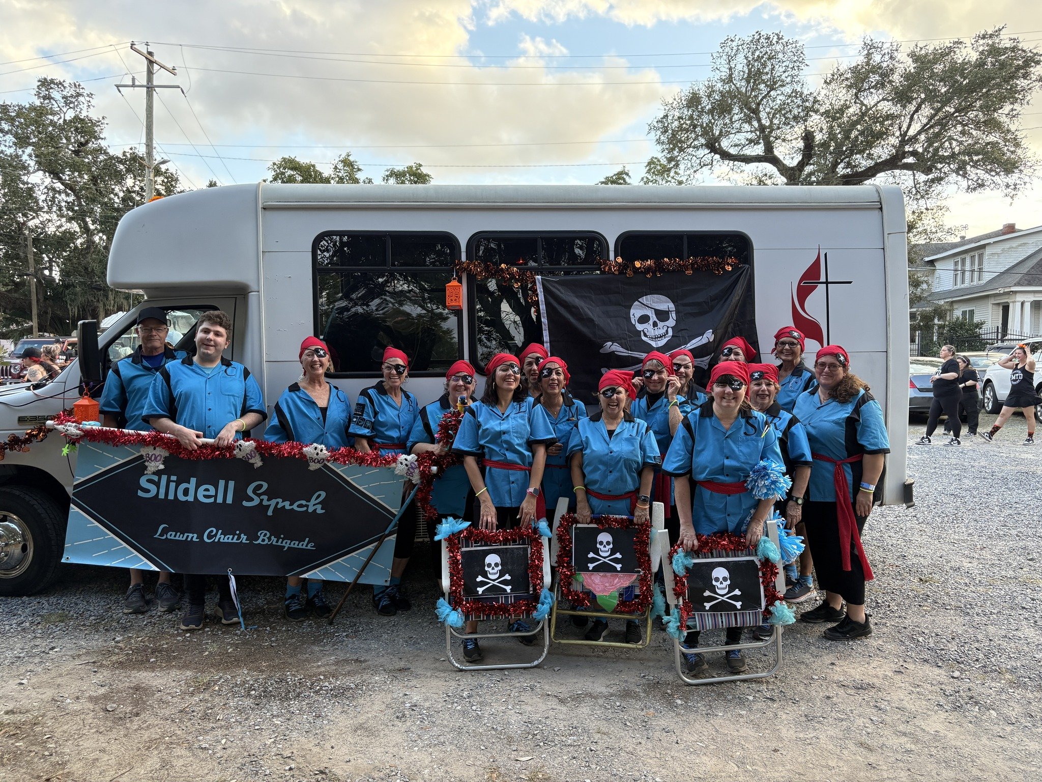 A group of people dressed in blue and red costumes, some wearing eye patches and hats, standing in front of a decorated float with pirate skull banners, during a parade or celebration.