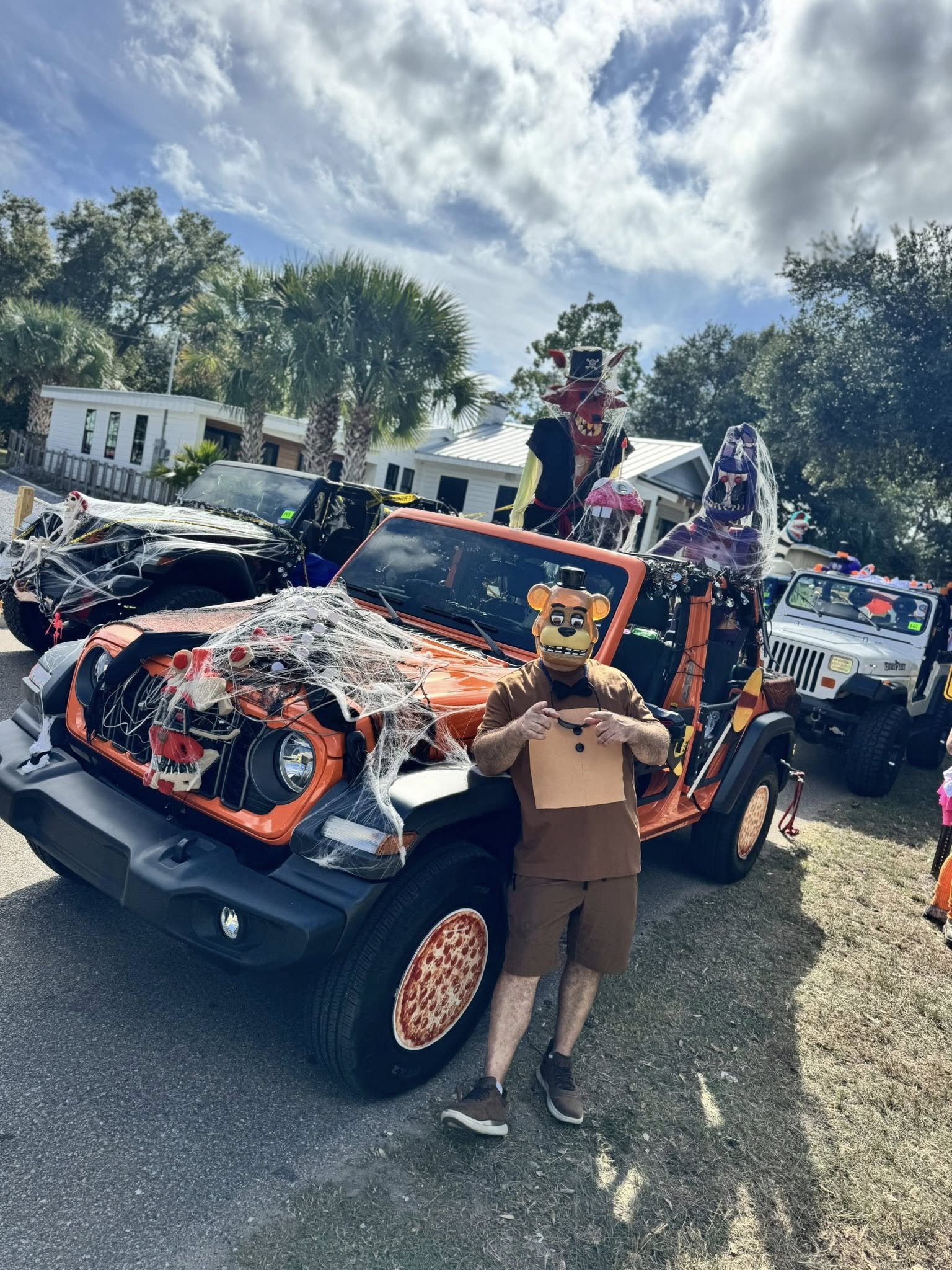 A person dressed in a scarecrow costume with a bear mask, standing in front of a decorated Jeep vehicle with Halloween decorations, including spider webs and Halloween-themed figures. The scene appears to be at a Halloween event outdoors on a sunny d