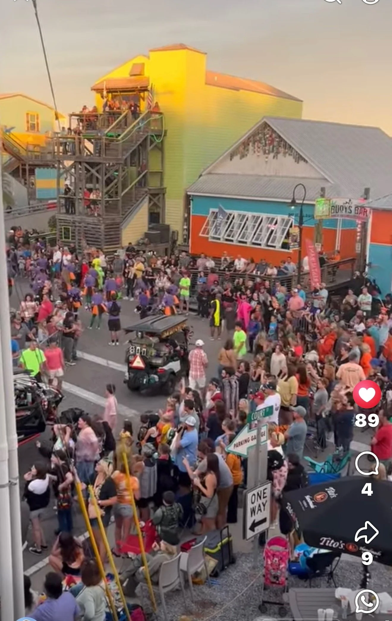 Crowded outdoor area with many people gathered, some sitting on benches, others walking or standing. Colorful buildings and signs, including one that says 'Buoys Bar' and another with an arrow pointing left. A small amusement ride with yellow poles a