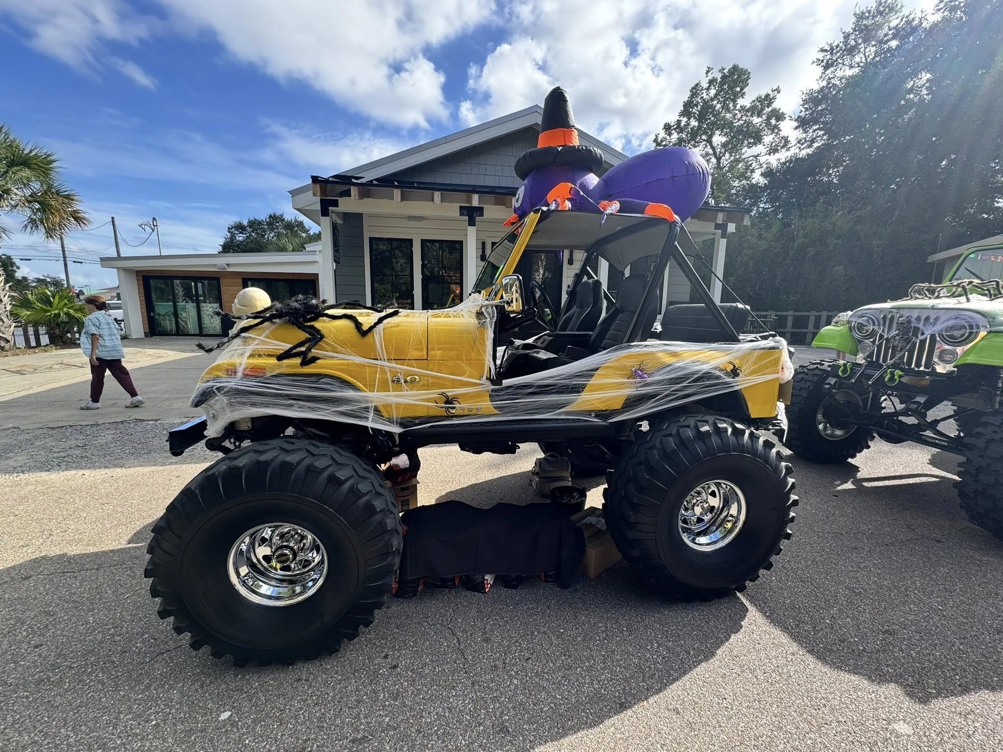 A yellow golf cart decorated for Halloween with fake cobwebs and spiders, parked outdoors in front of a modern house with a gray roof and large windows. There are Halloween decorations on the roof of the cart, including a purple witch hat and a purpl