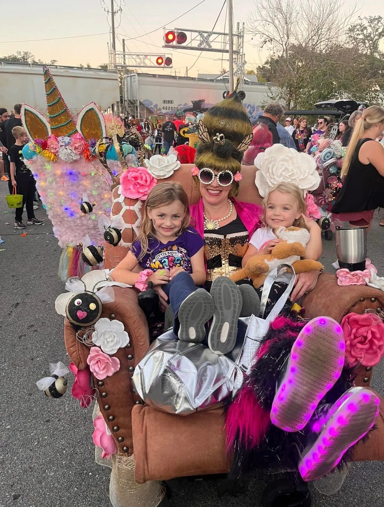 Woman dressed as a fairy tale character with two young girls sitting on her lap, all smiling. They are outdoors at a festive event, sitting on a pink armchair decorated with flowers, bees, and fairy wings. The woman wears a large flower headpiece, be