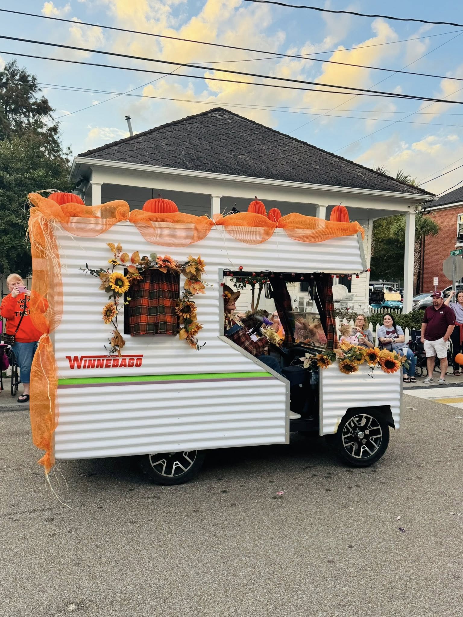 A small decorated trailer with orange drapes, sunflowers, and plaid fabric, parked on a street during a parade, with people watching and participating in the event, houses and trees in the background, and a clear sky overhead.
