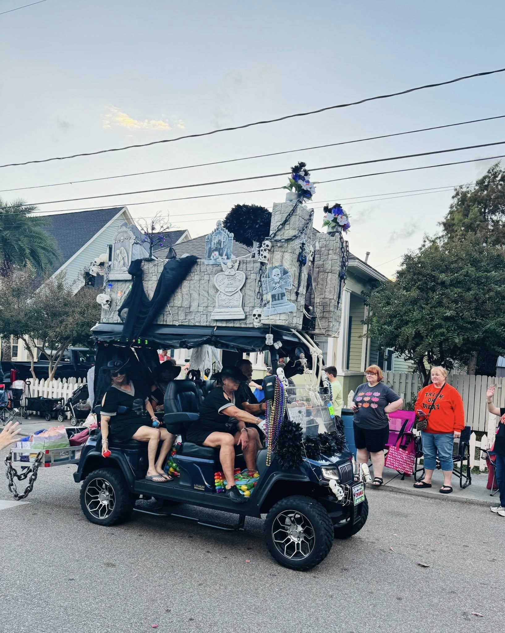 A black golf cart decorated with Halloween-themed items, including skulls and skeletons, is parked on a street during a parade or festival. There are people in costume or casual clothing around, some sitting on the golf cart and others standing nearb
