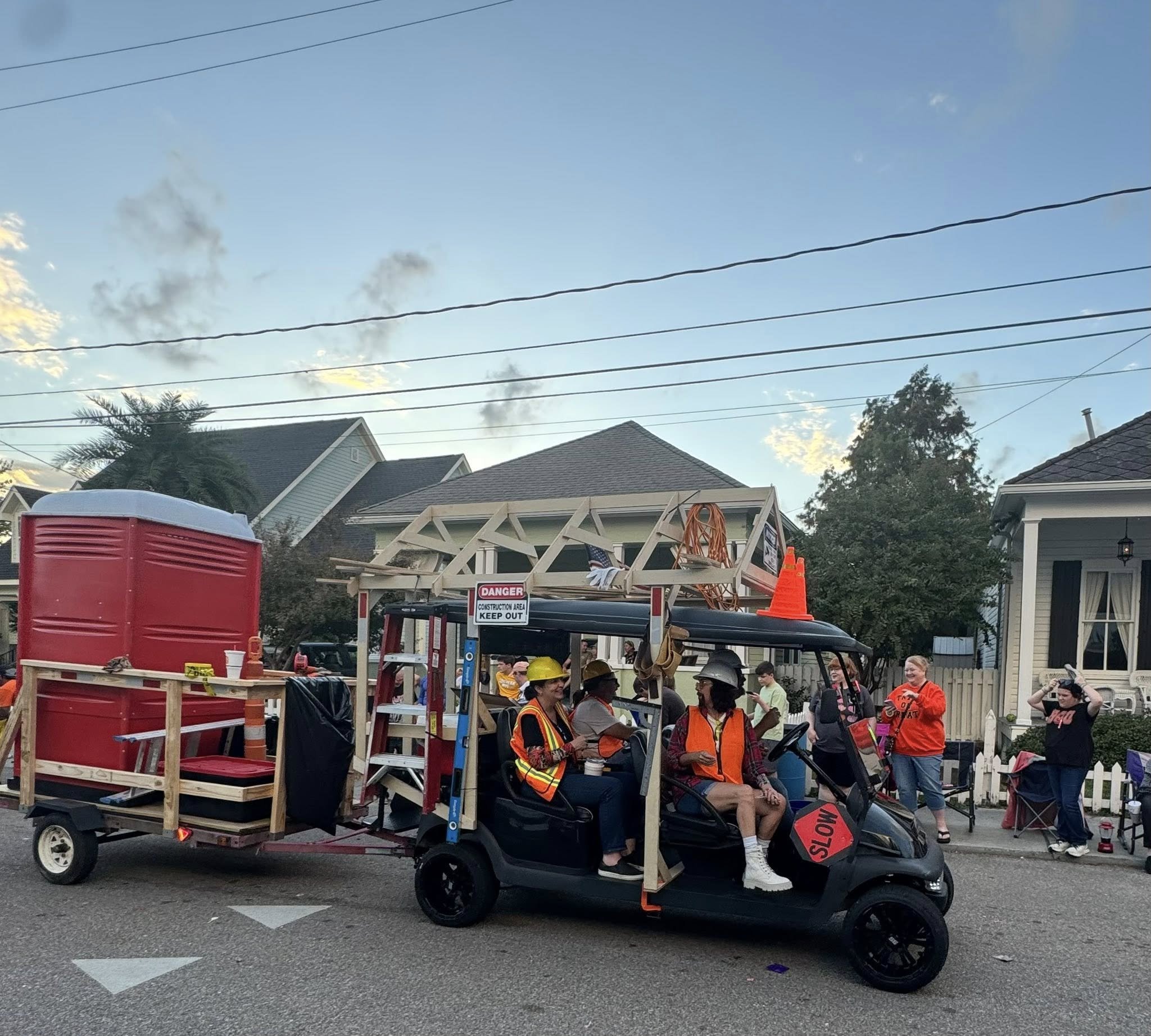 A golf cart decorated as a parade float with people in safety vests and hats, some sitting and some standing, in a neighborhood street with houses and trees in the background. The float has a wooden framework with a sign that says "DANGER", orange co
