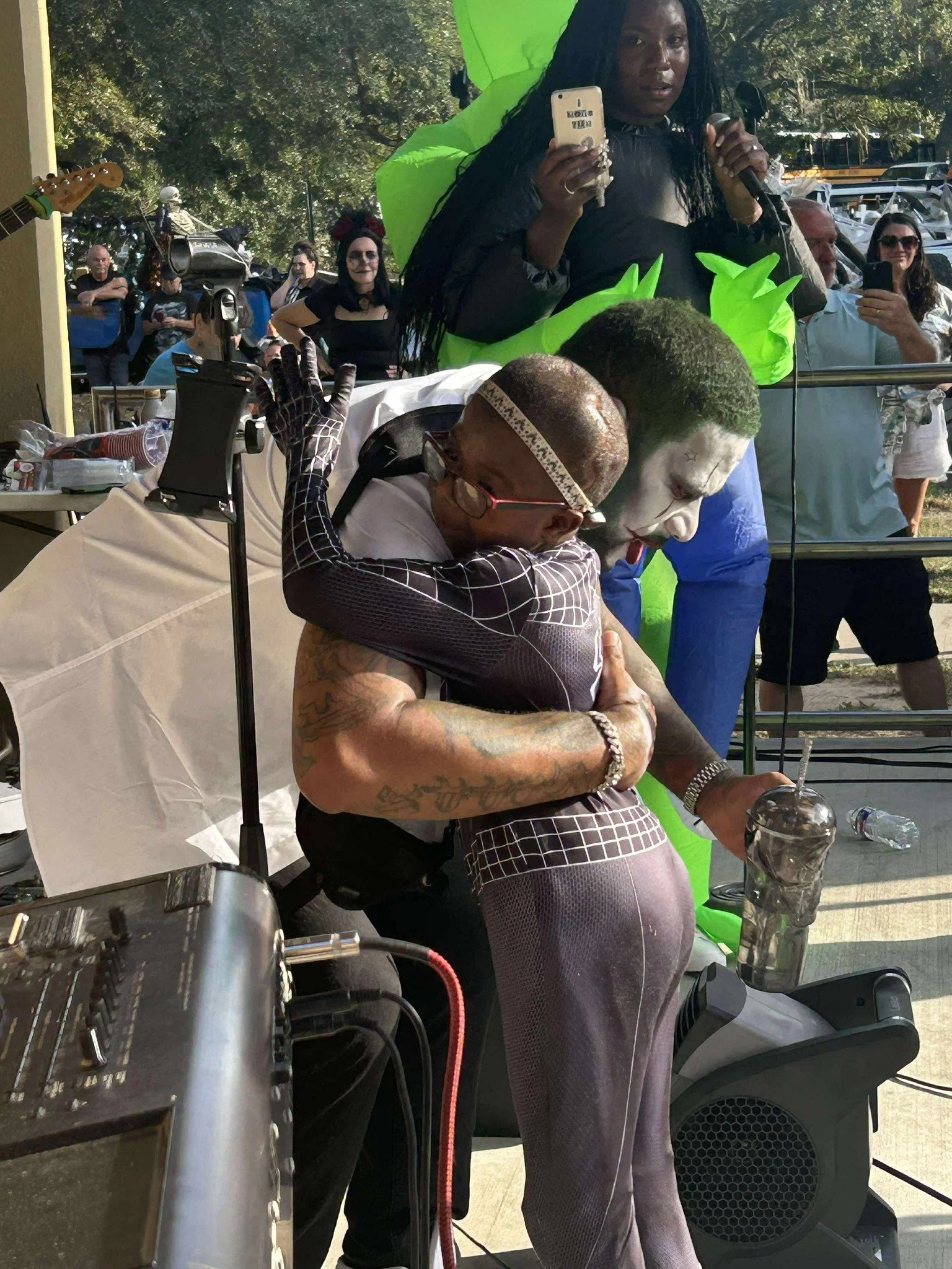 A woman with long braids and dark skin, holding a smartphone and a microphone, standing in front of a mirror during an outdoor event. She is dressed in black and green with a neon green jacket. In front of her, a man with a shaved head, glasses, tatt