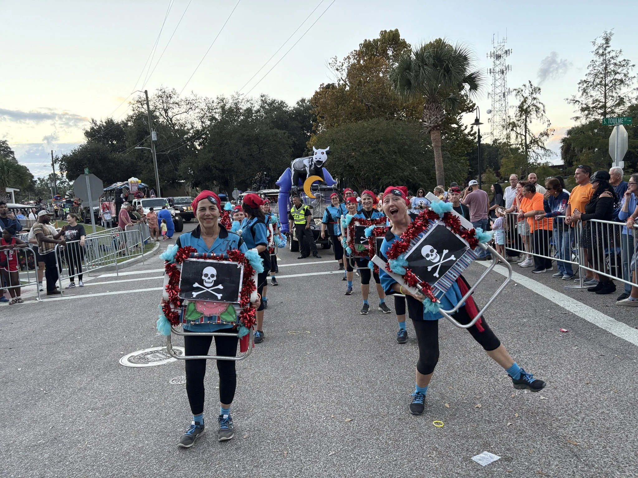 Group of people participating in a parade wearing costumes themed around pirates and skulls, walking along a street lined with onlookers, trees, and utility poles, with a large inflatable zebra and a float in the background.